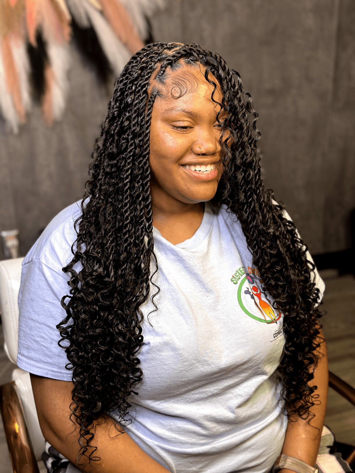 Young woman with long, curly hair smiling, wearing a light-colored T-shirt, in front of a textured background.