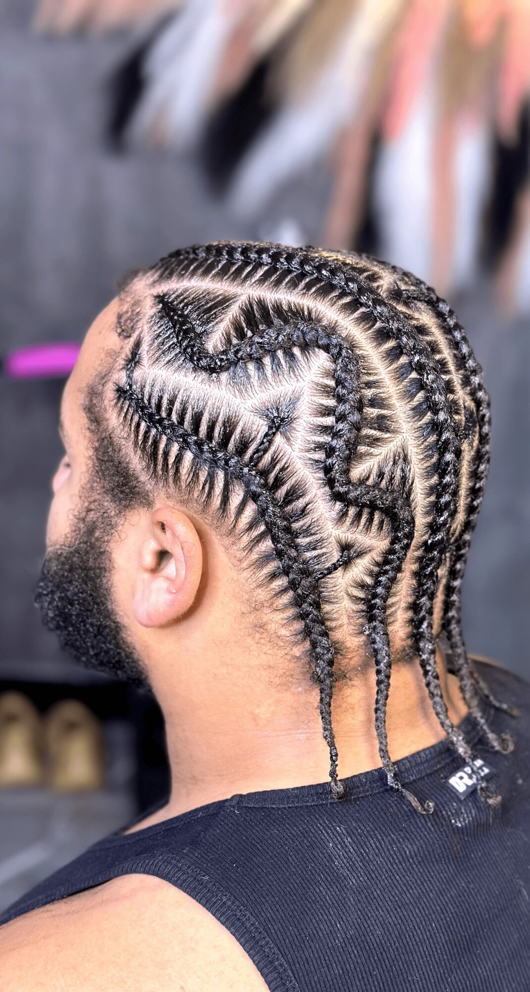 Side view of a man with intricate cornrow hairstyle, featuring zigzag and curved patterns, with a beard and black sleeveless shirt.
