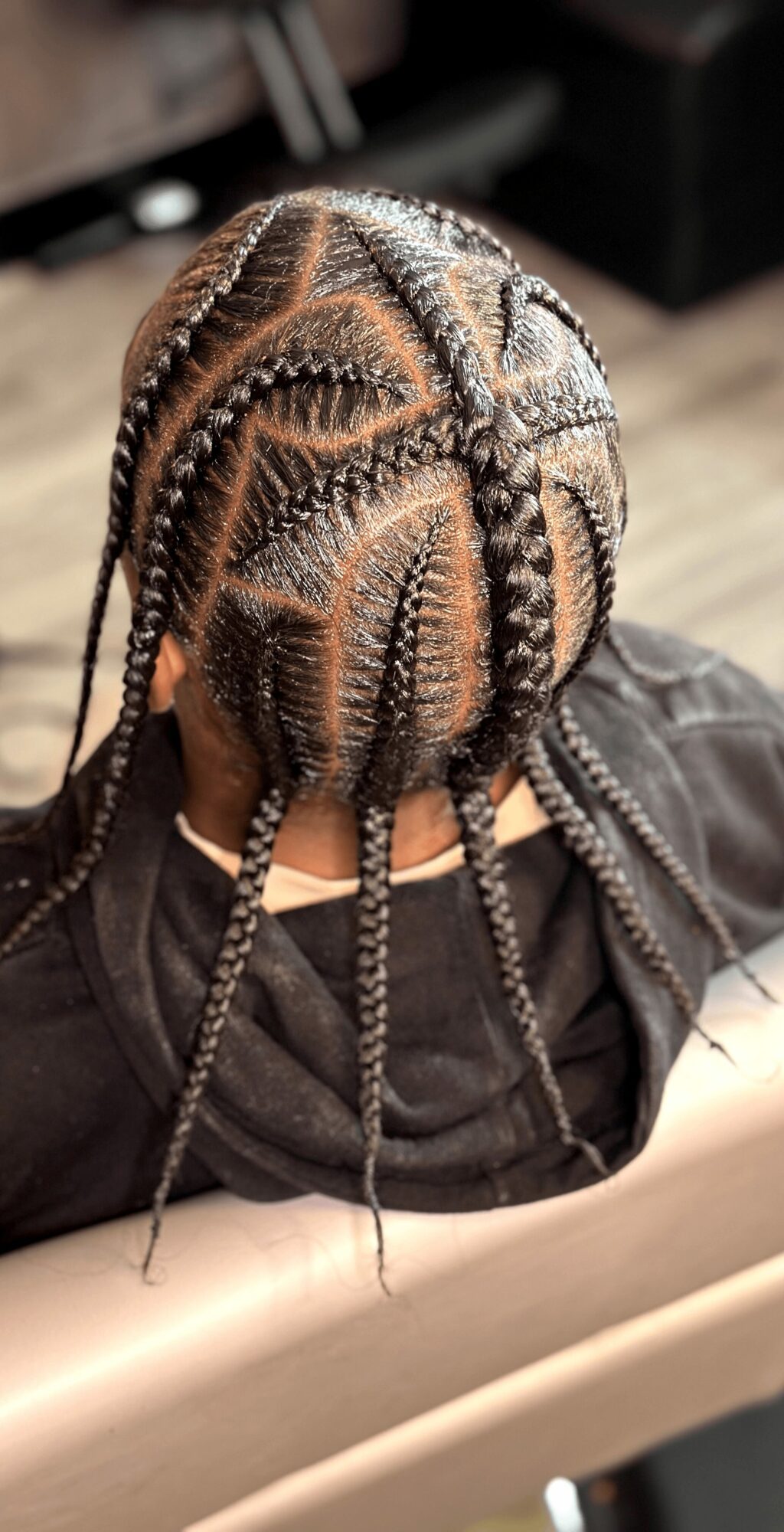 Top view of a person's head with braided hair styled in intricate patterns, wearing a dark shirt, sitting on a beige surface.