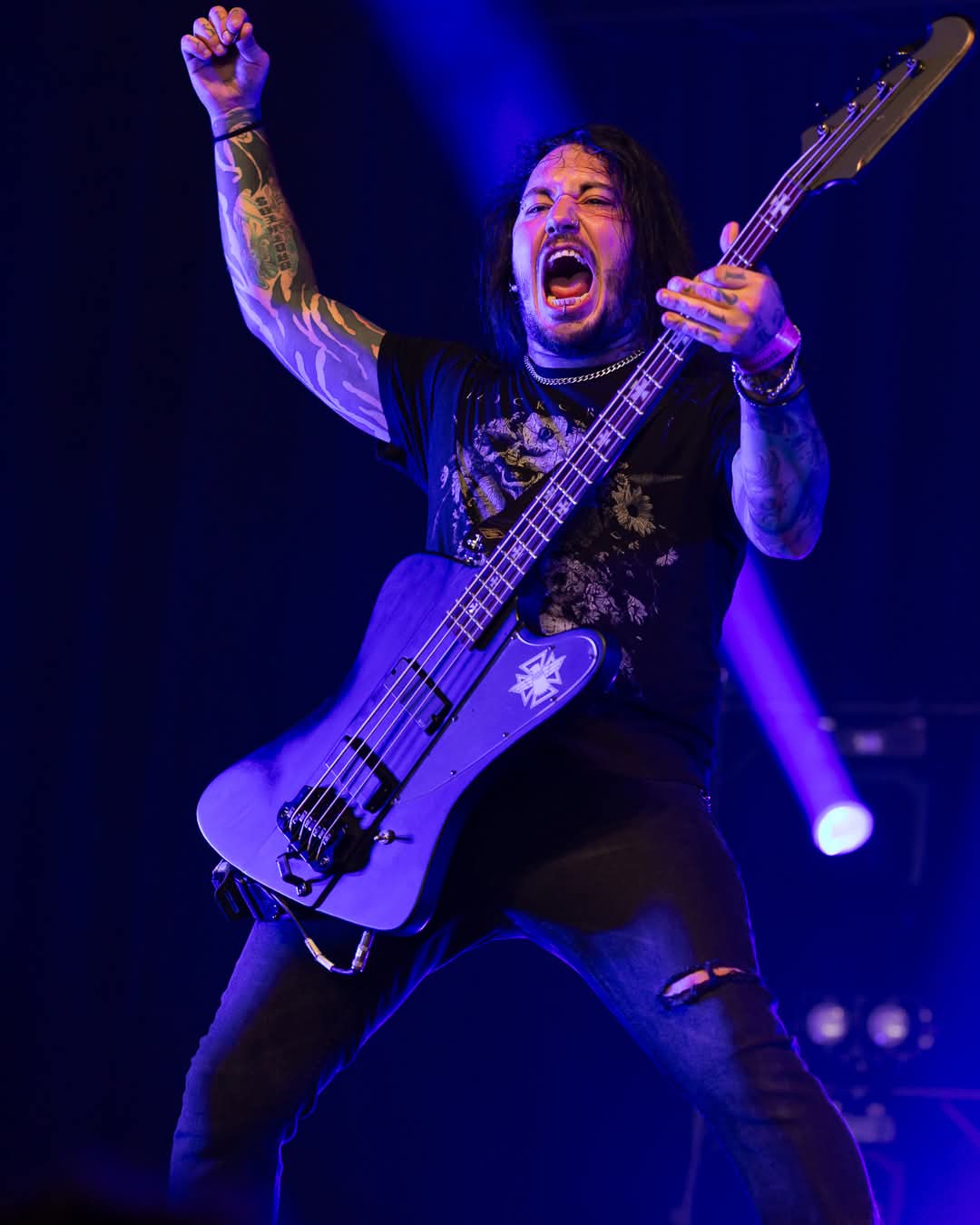 Musician playing guitar on stage, raising one arm, with long hair and tattoos, under blue stage lighting.