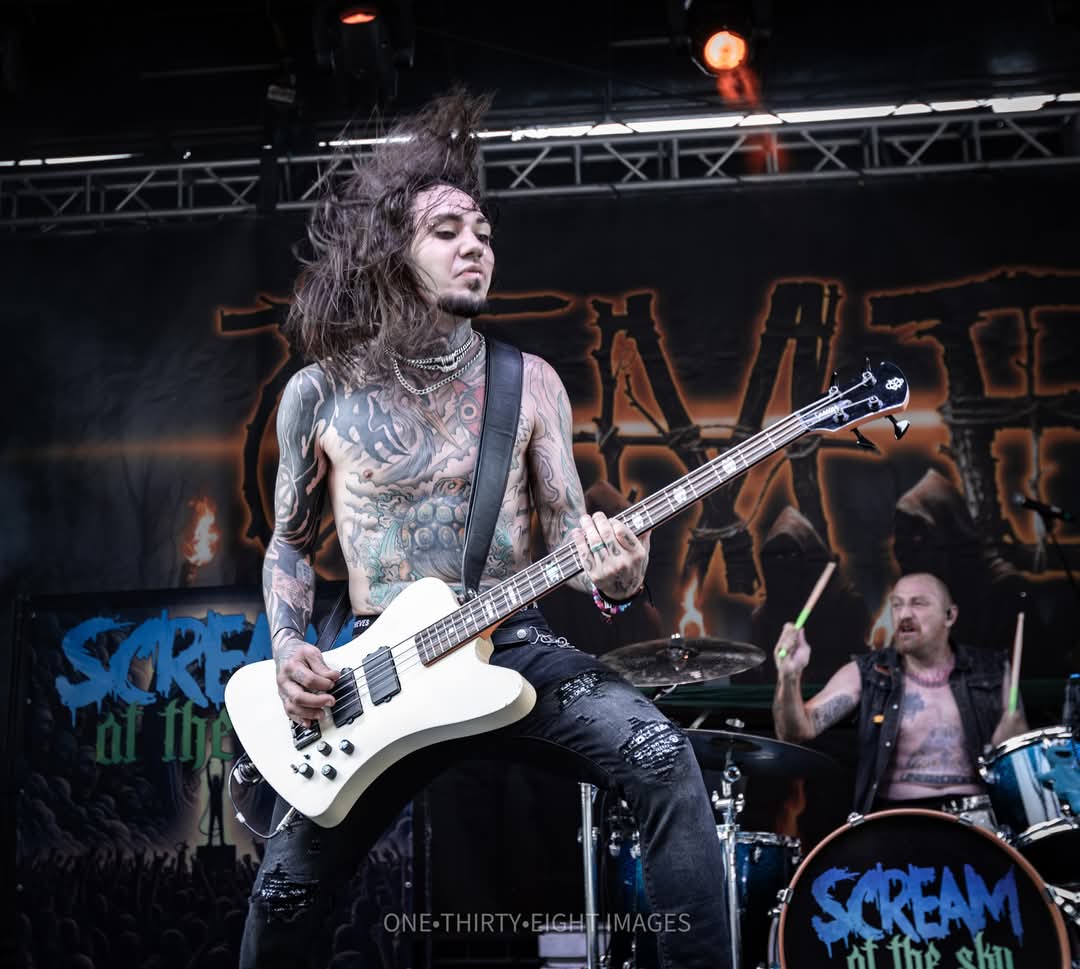 Musician with long hair playing an electric guitar on stage during a concert, with a drummer in the background.