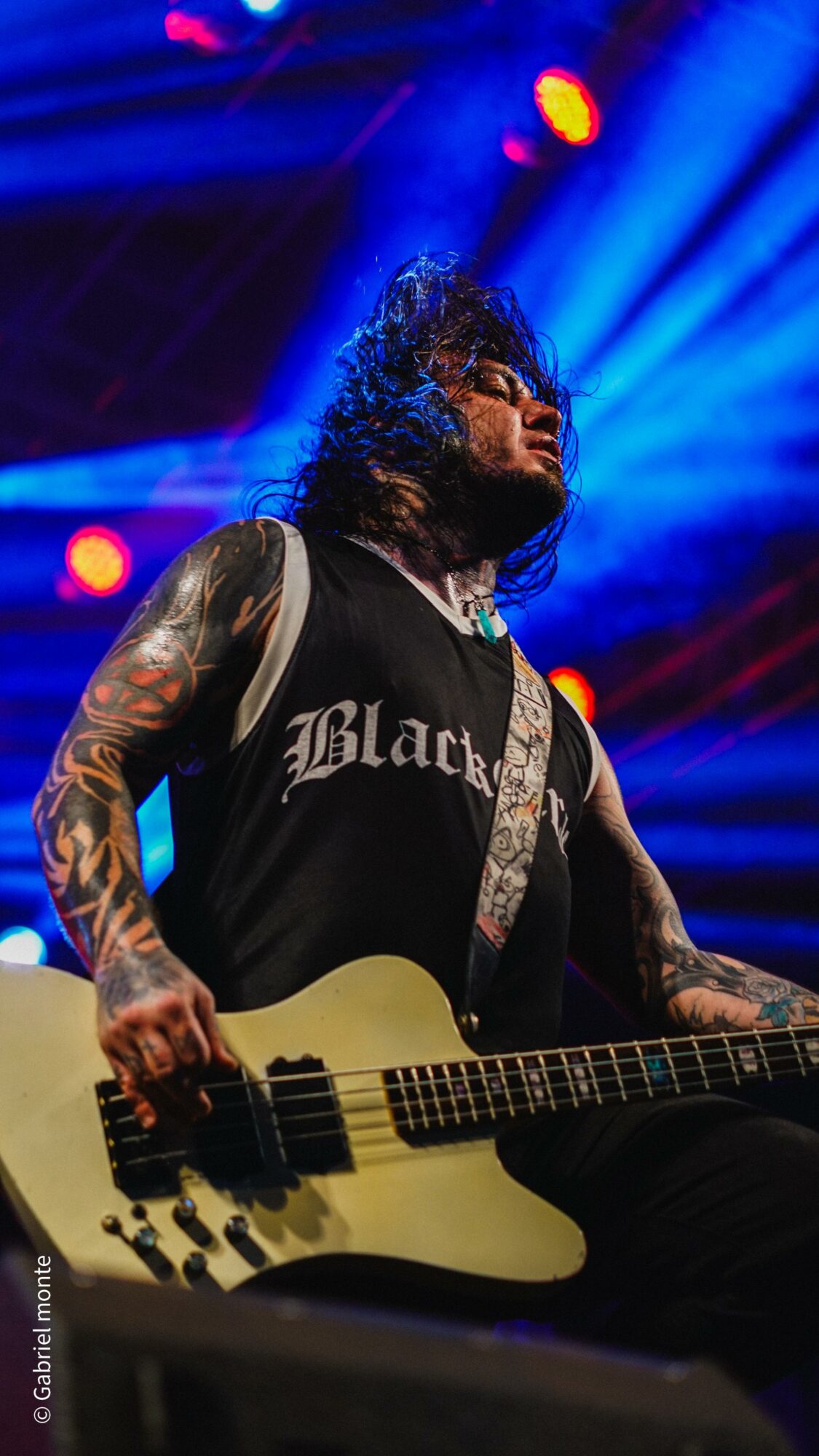 Musician with tattoos playing guitar on stage with blue and red lights, wearing a black sleeveless shirt.