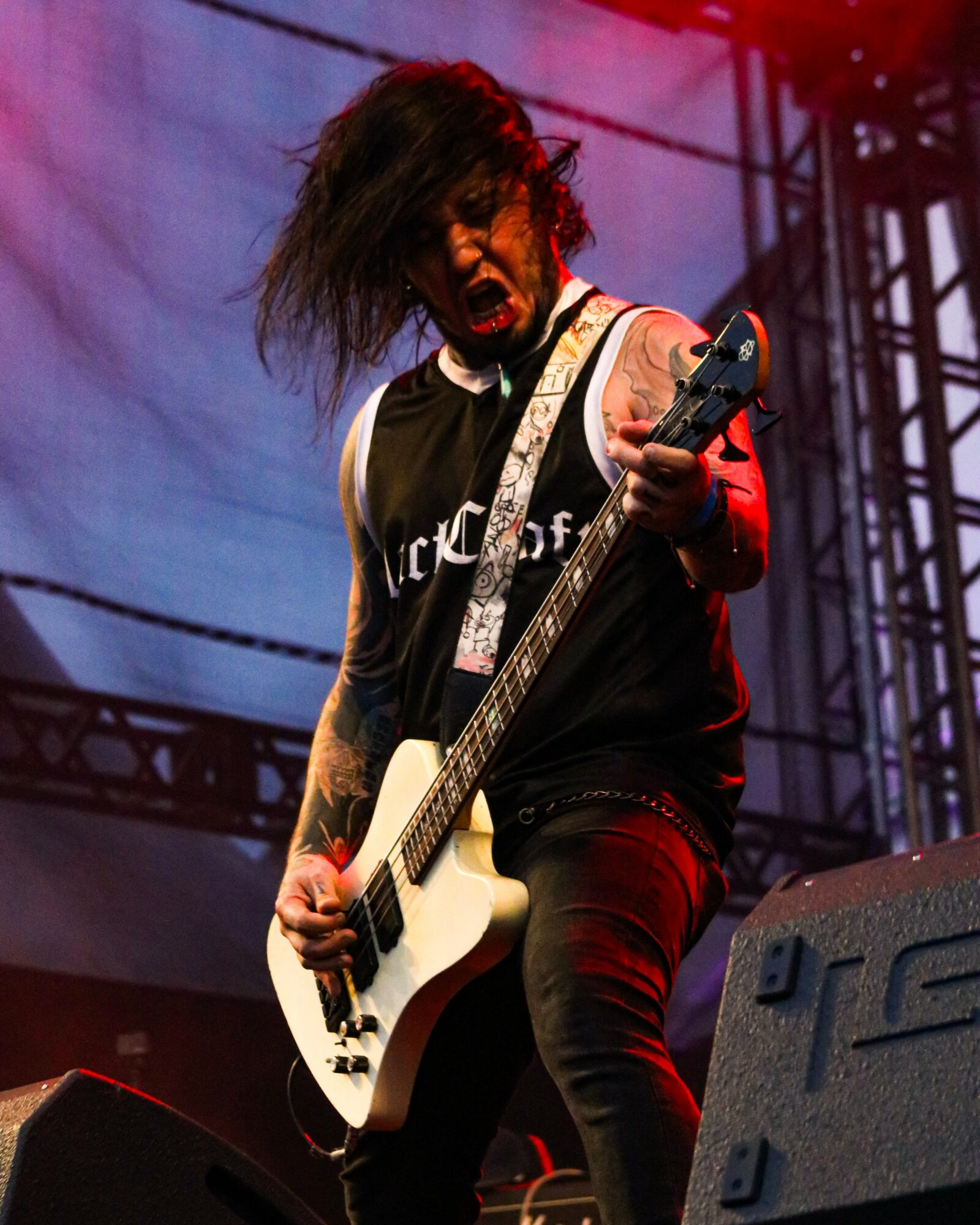 Musician with messy hair playing a white electric guitar on stage, expressive face, wearing a black sleeveless shirt, stage lighting in background.