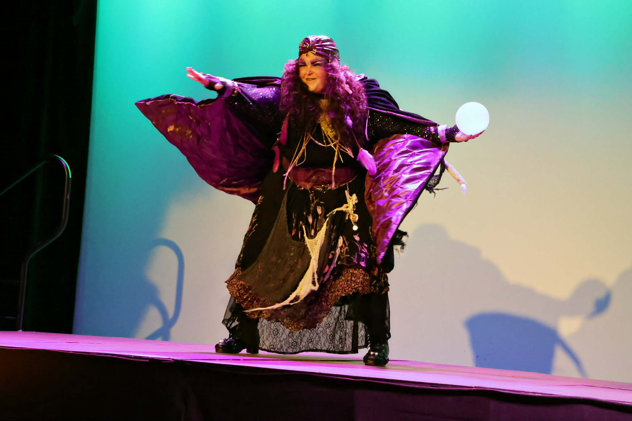 Performer in colorful costume on stage with arms outstretched, holding a ball, with a gradient background.