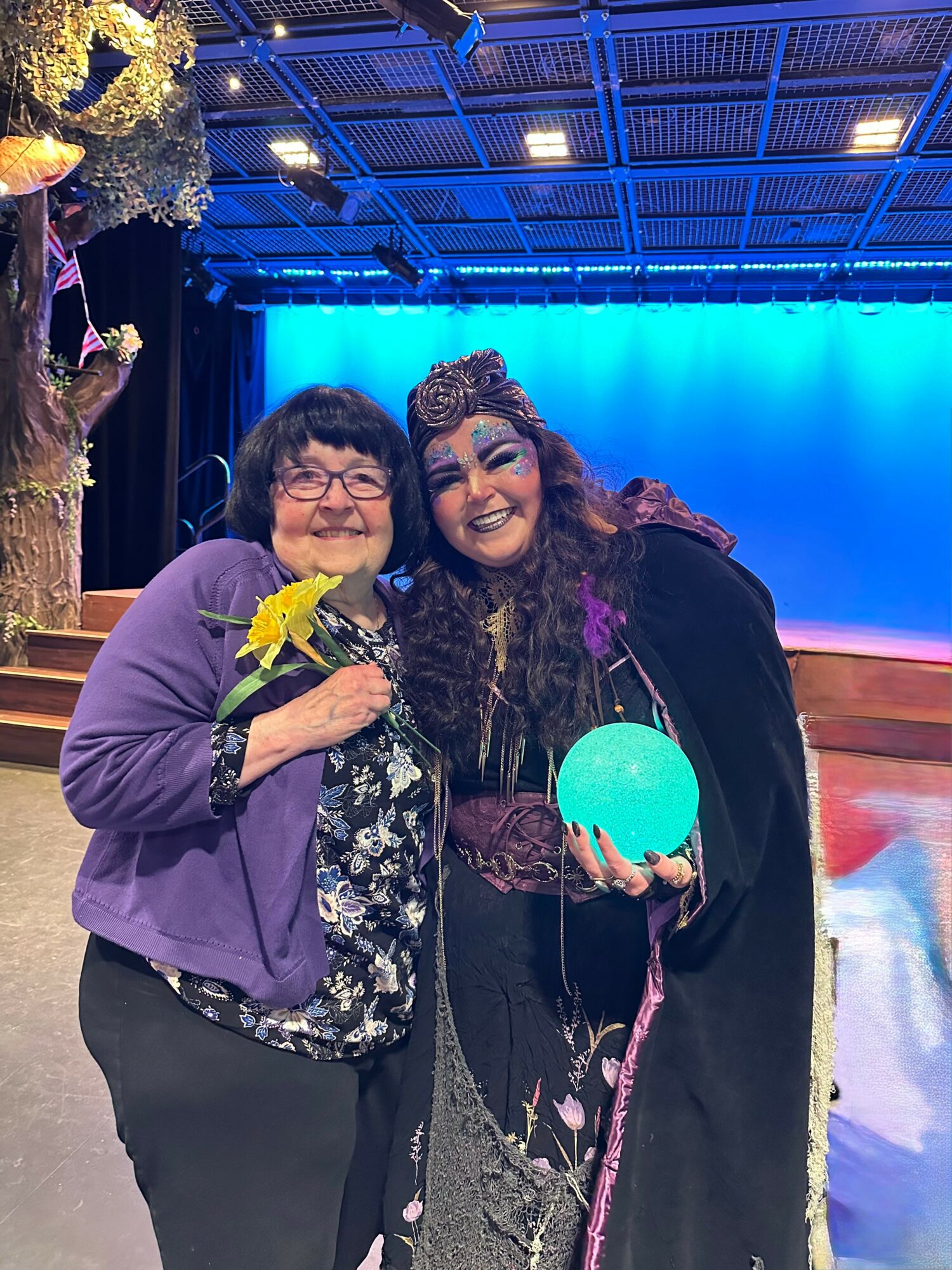 Two women smiling on stage, one holding a yellow flower, with a blue background and stage lights.