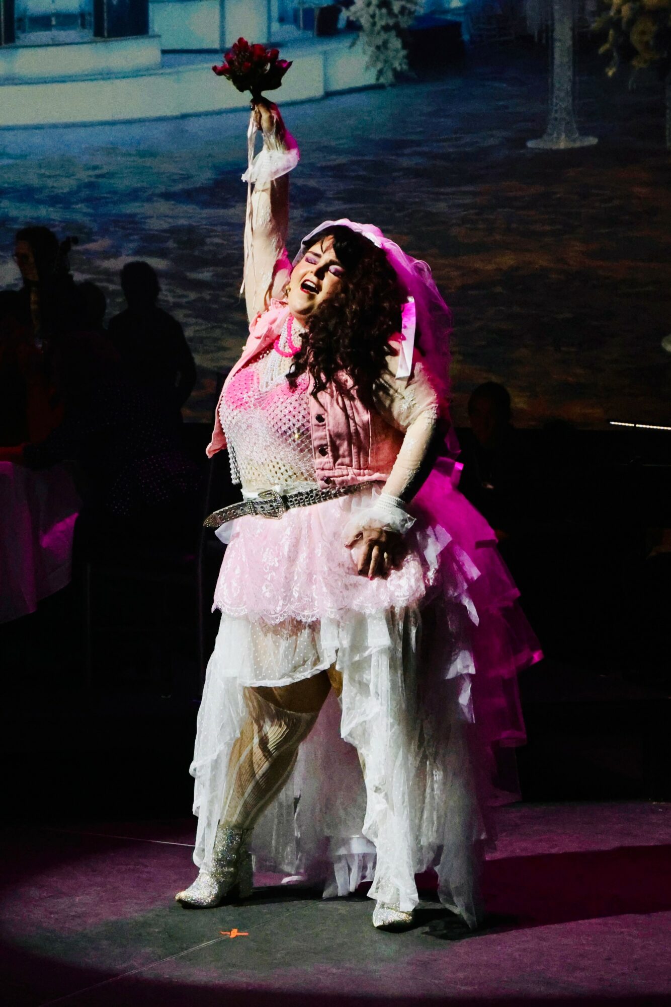Performer on stage holding a bouquet of flowers, wearing a pink and white costume with ruffled layers, smiling.