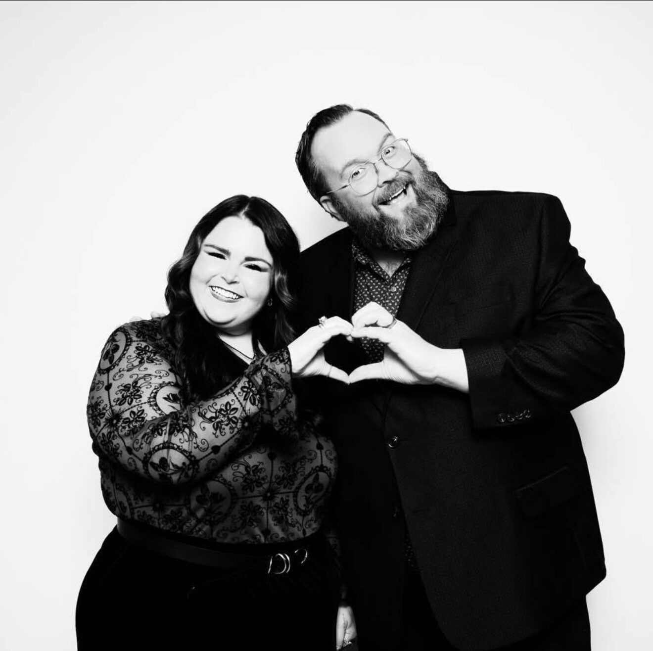Two people smiling, forming a heart shape with their hands, black and white photo, both wearing dark clothing.