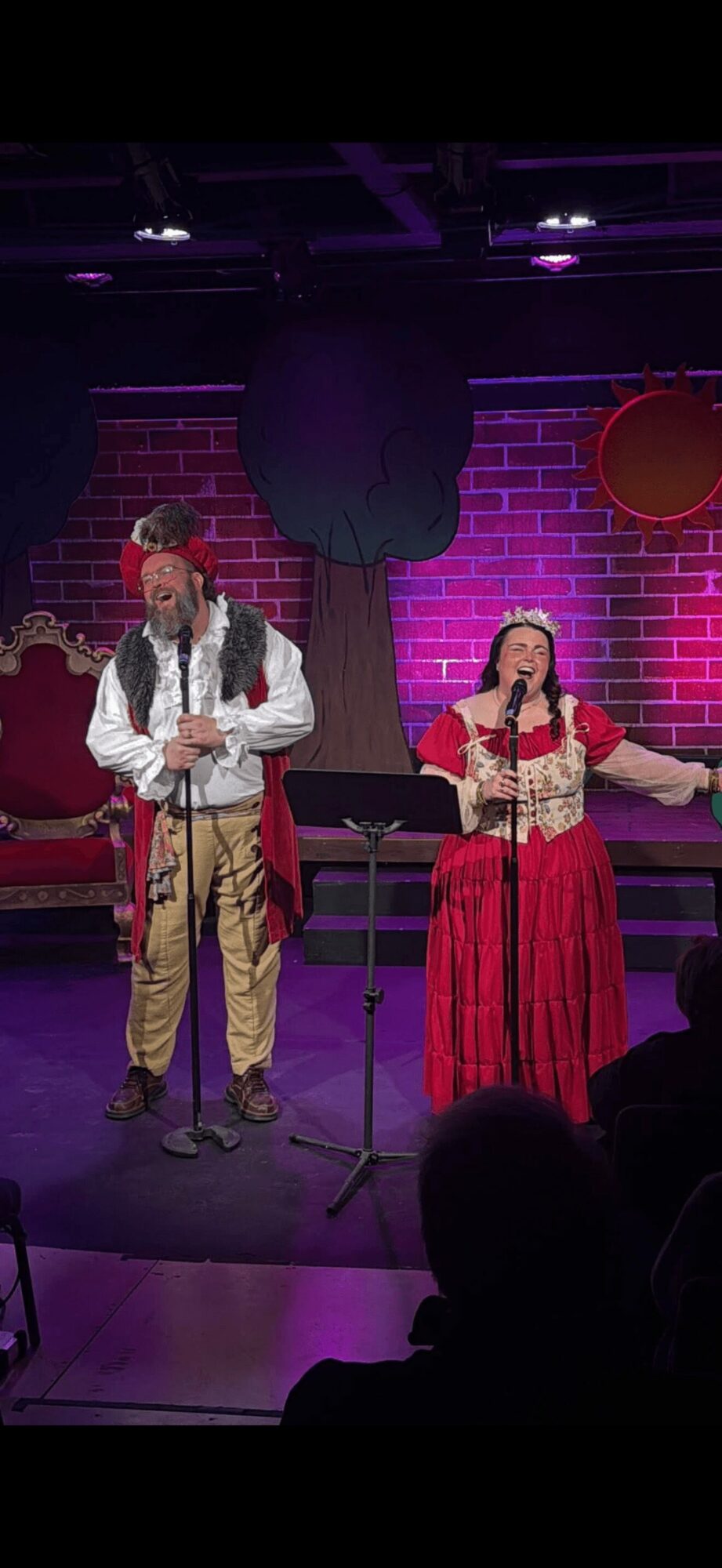 Two performers on stage with a brick wall background, one in traditional costume, singing into microphones.