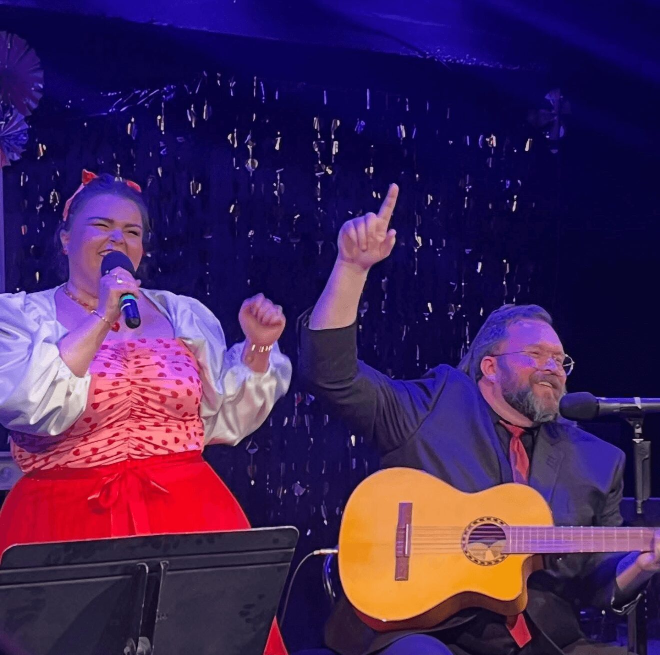 A woman singing into a microphone and a man playing guitar on stage with a dark, sparkly background.