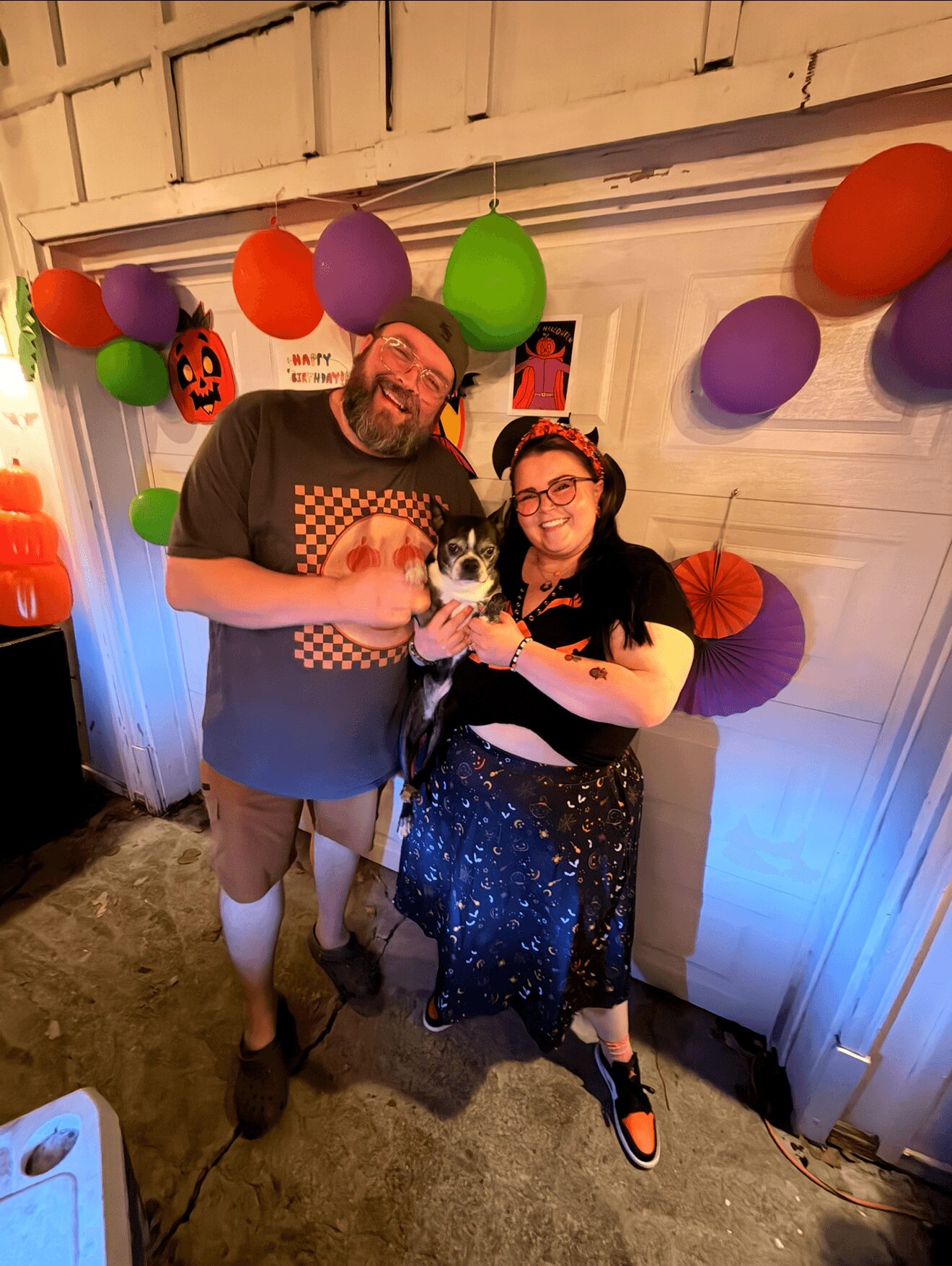 Two people, a man and a woman, stand together at a party with colorful balloons and decorations behind them. The man has a beard and glasses, wearing a dark t-shirt and shorts. The woman has glasses, dark hair, and is wearing a black dress with a floral pattern, holding a small dog. They are smiling and posing for the photo.
