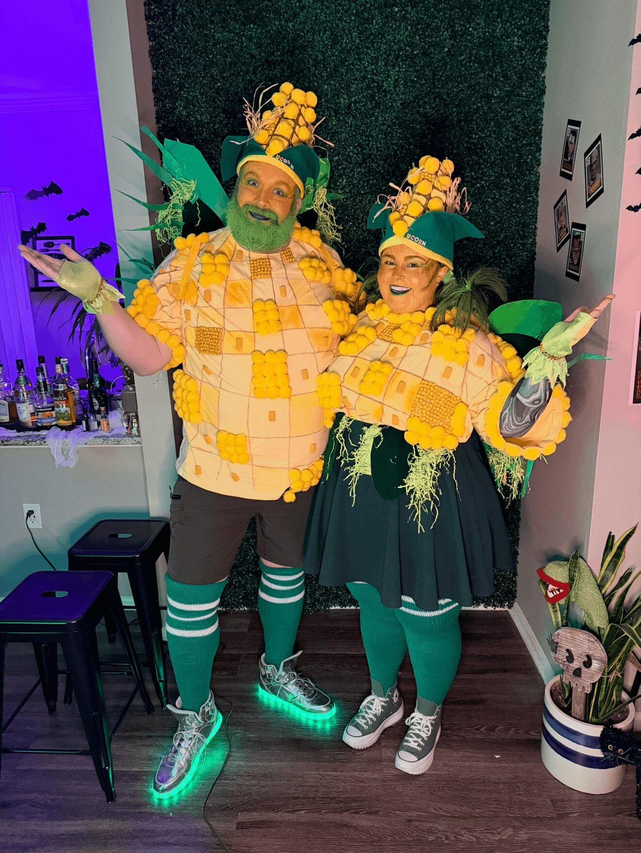 Two people in pineapple-themed costumes pose indoors, smiling with arms outstretched, surrounded by plants and decorations.