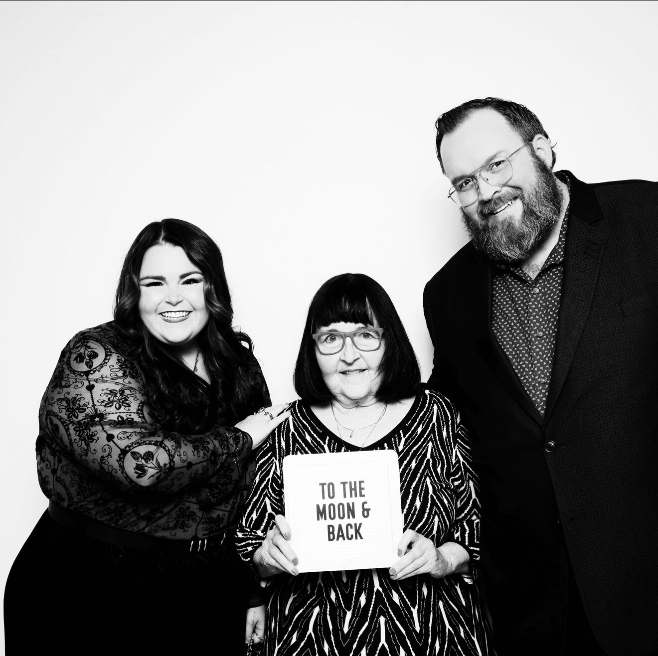 Three people smiling, one holding a sign that says 'To the Moon & Back', against a plain background.