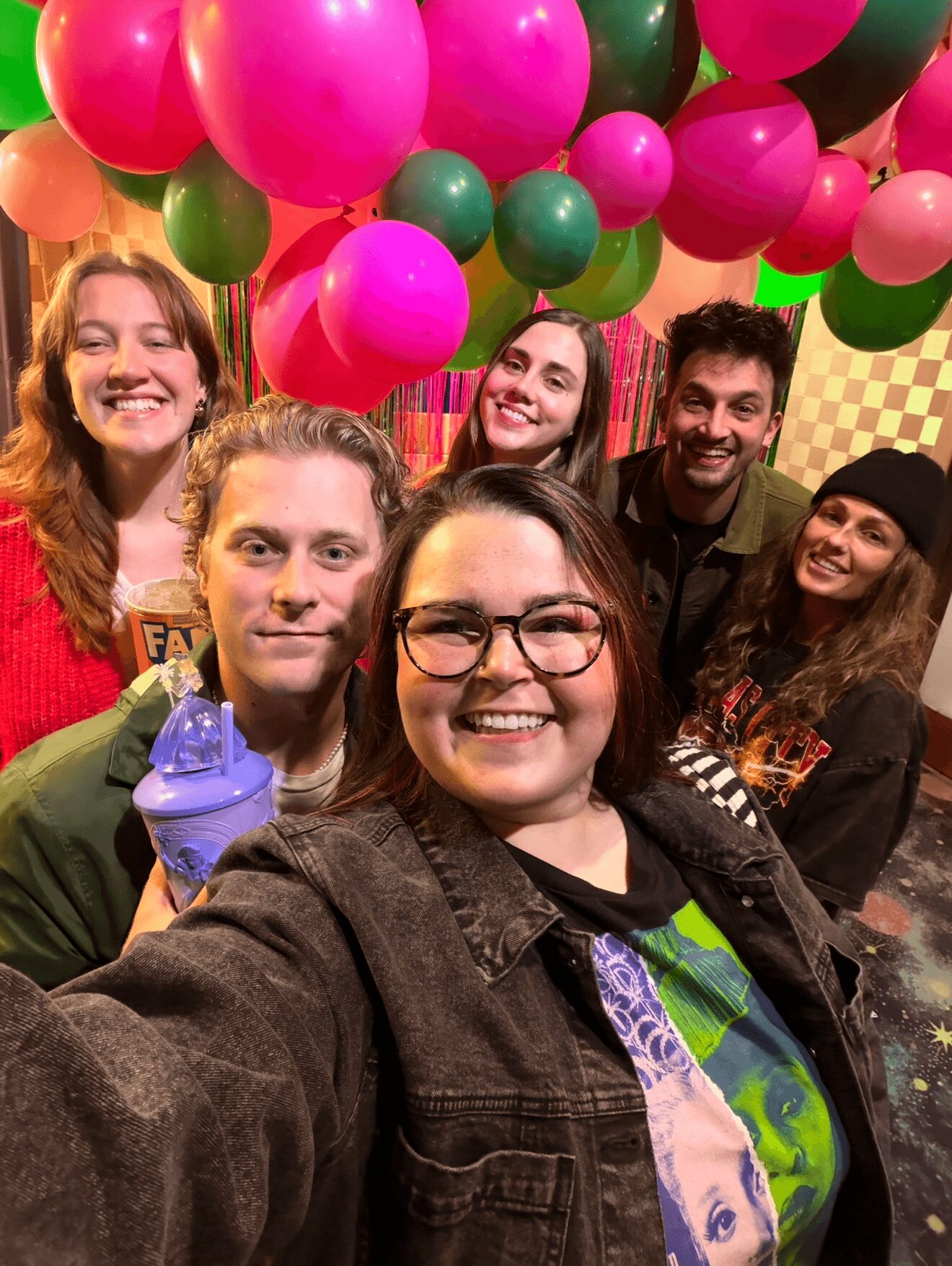 Group of six smiling people taking a selfie with colorful balloons overhead in a festive setting.