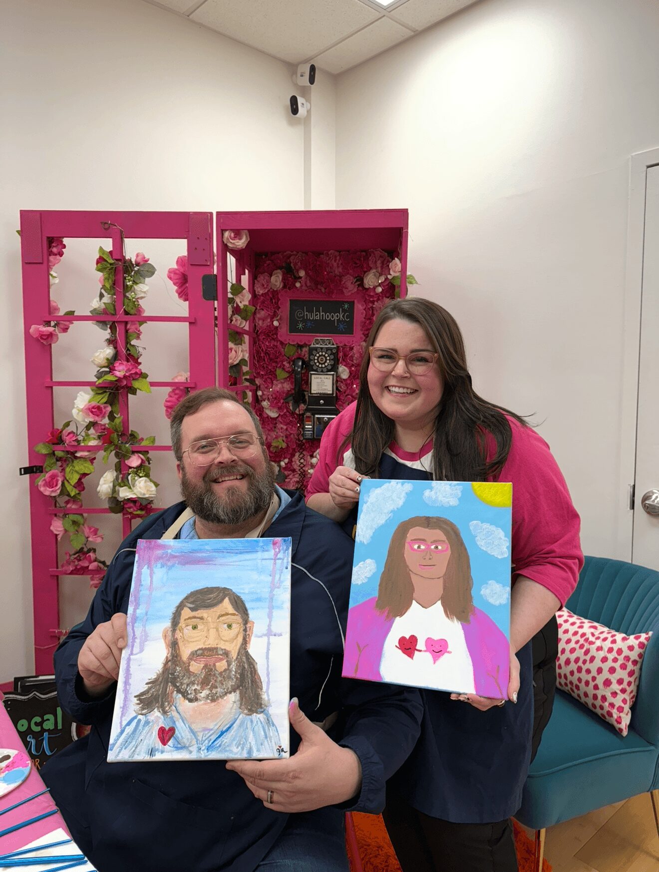 Two people smiling and holding colorful portraits of themselves, with a pink floral display in the background.