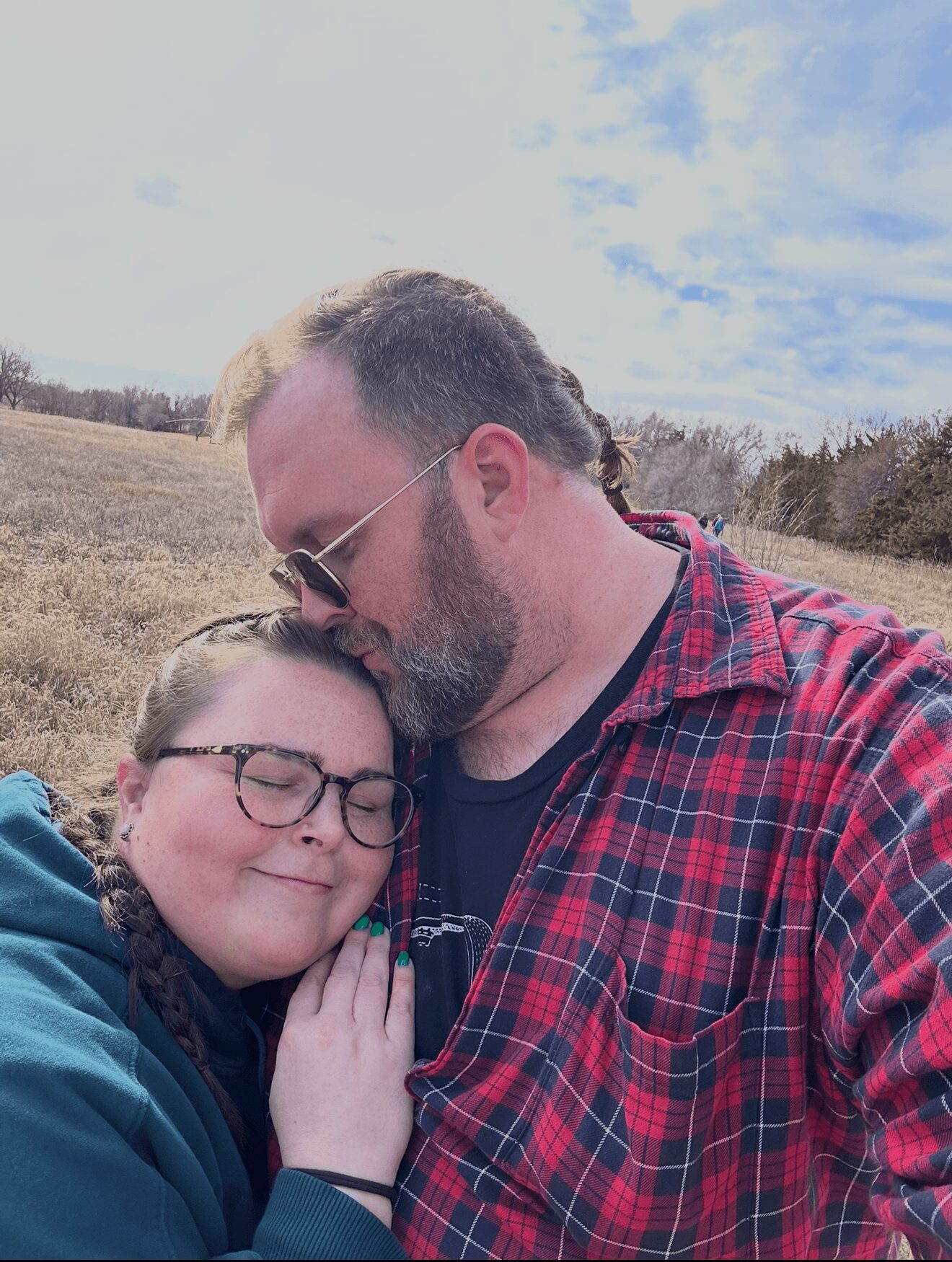 A man and a girl are outdoors, hugging with eyes closed, smiling, in a field with a partly cloudy sky.