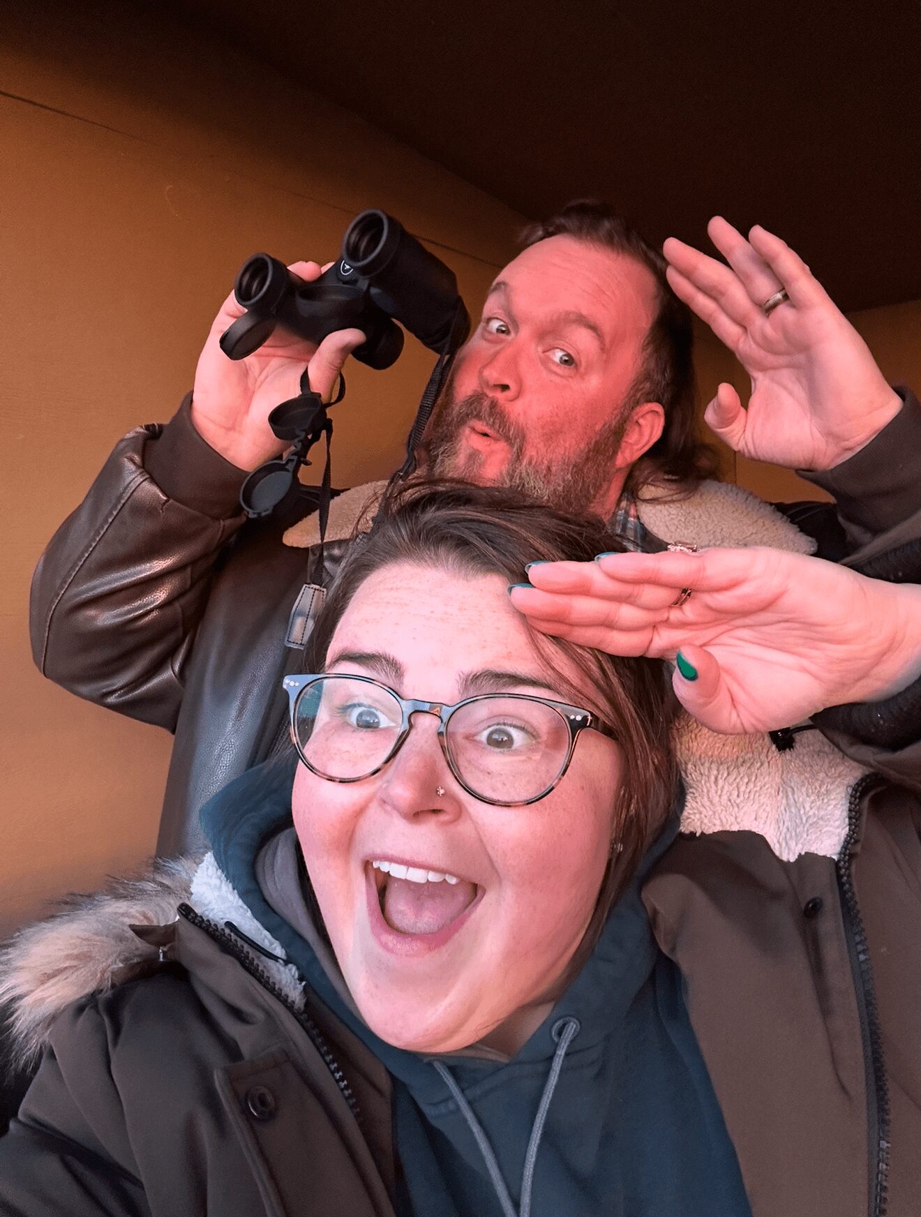 Two people taking a selfie, one holding binoculars, both smiling and making playful gestures, indoors with a brown background.