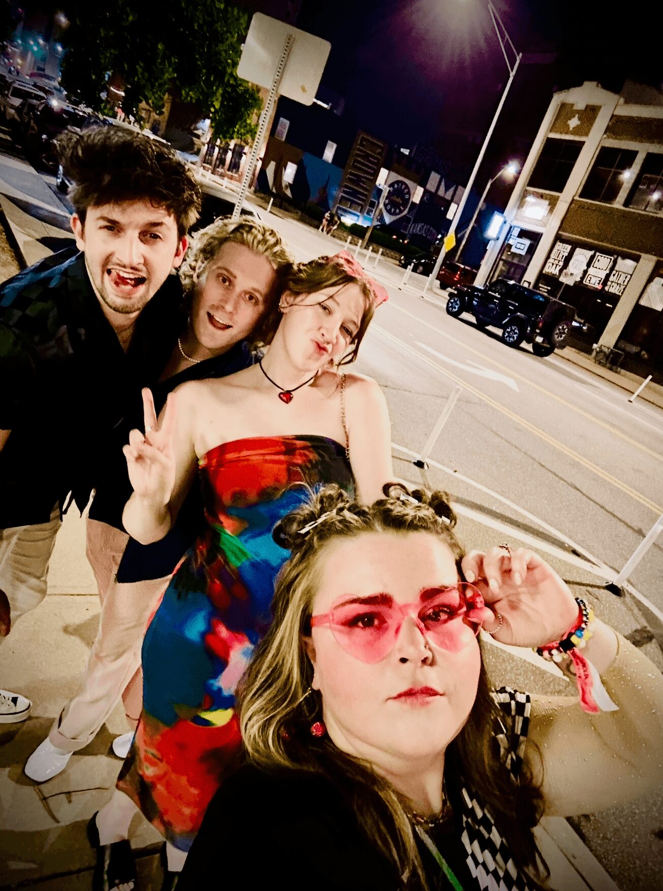 Four young people posing outdoors at night, one making a peace sign, in front of a street scene with buildings and cars.