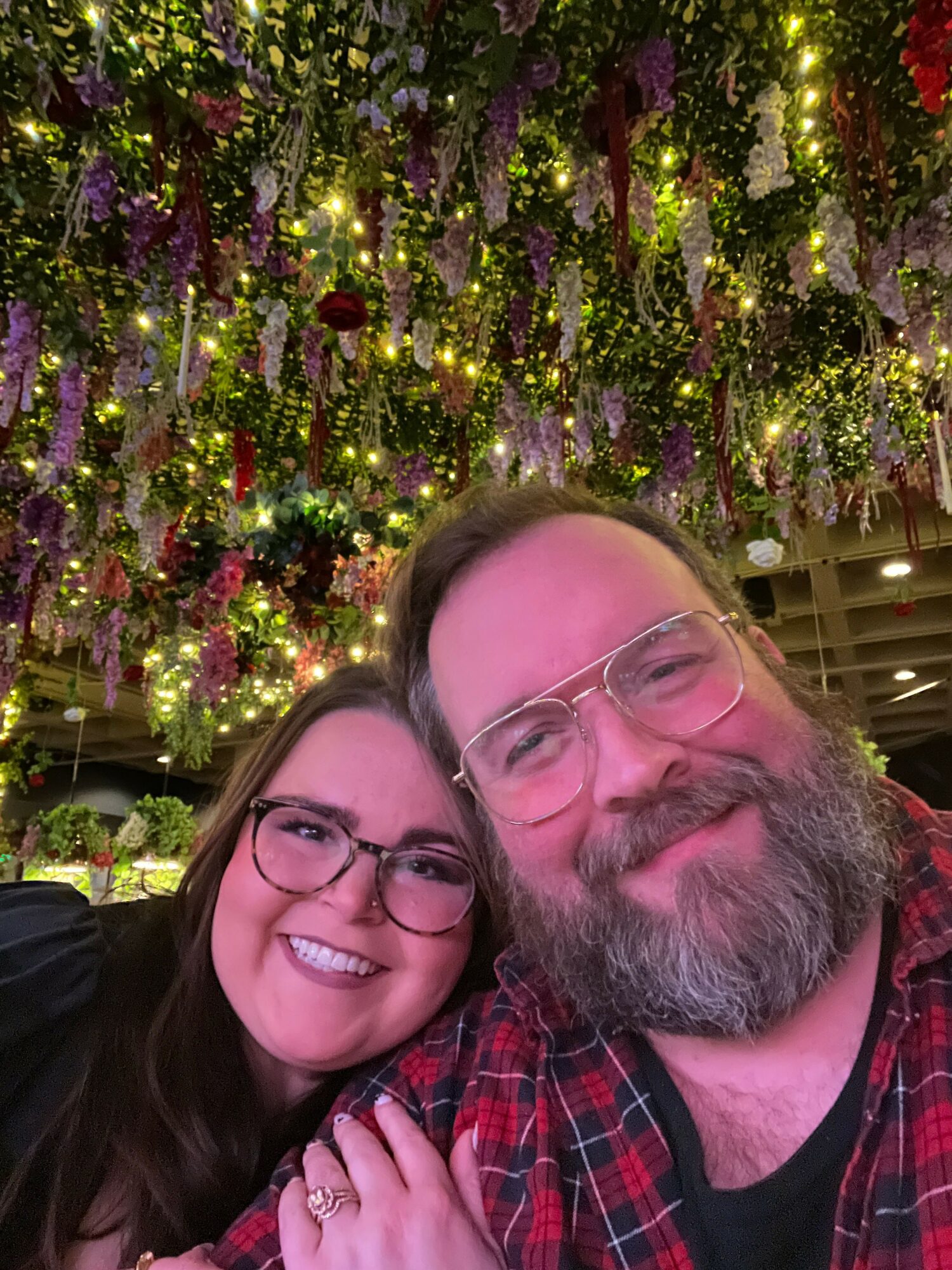 Two people smiling, wearing glasses, close together, in front of decorated Christmas trees with lights and ornaments.