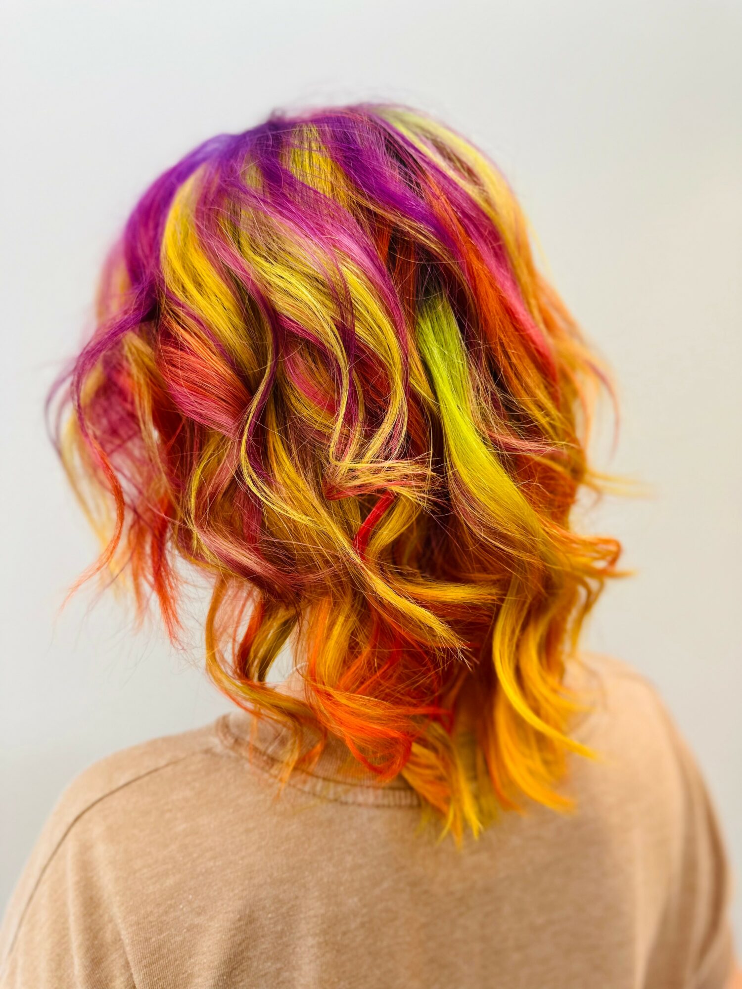 Back view of a person with colorful, wavy hair in pink, yellow, orange, and green shades, wearing a beige top.