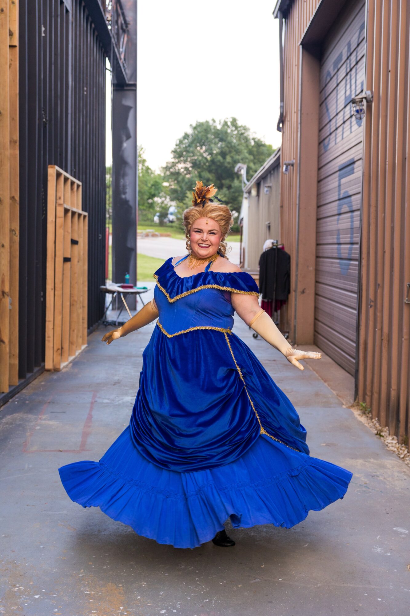 Woman in a blue dress twirling in an alleyway between wooden buildings, smiling, with trees and sky in the background.