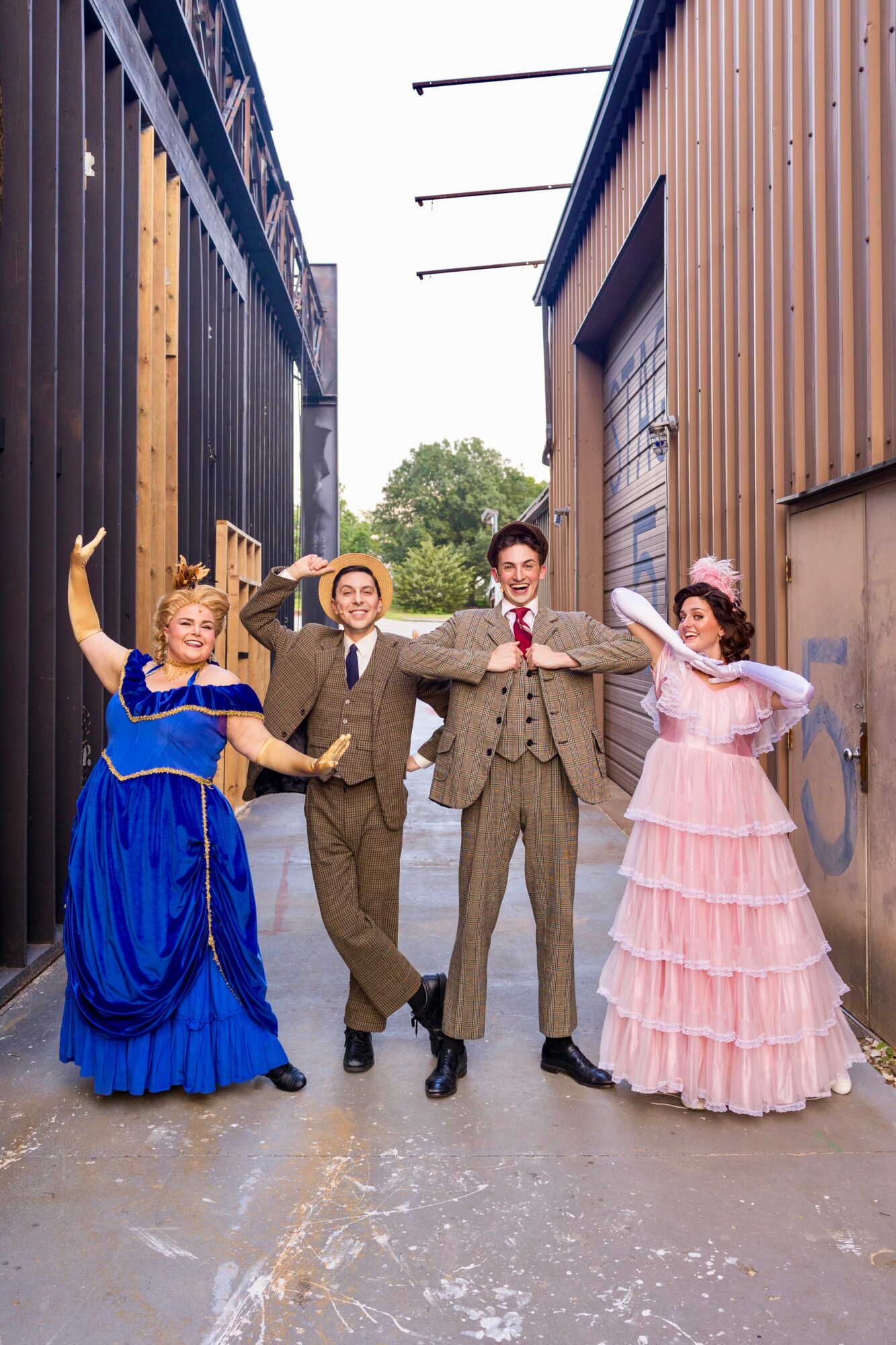 Four people in vintage costumes pose between two buildings, smiling and holding hands, with a cloudy sky above.