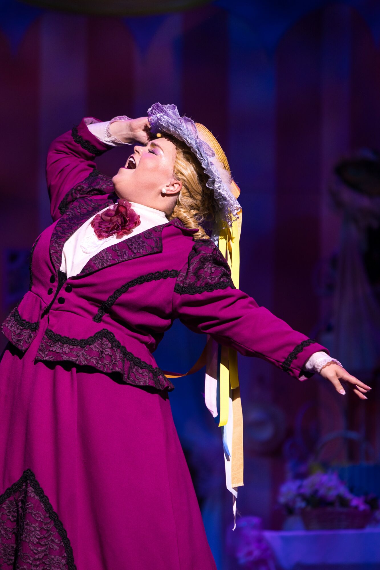 Performer in colorful costume with hat, arms outstretched, on stage with purple backdrop.