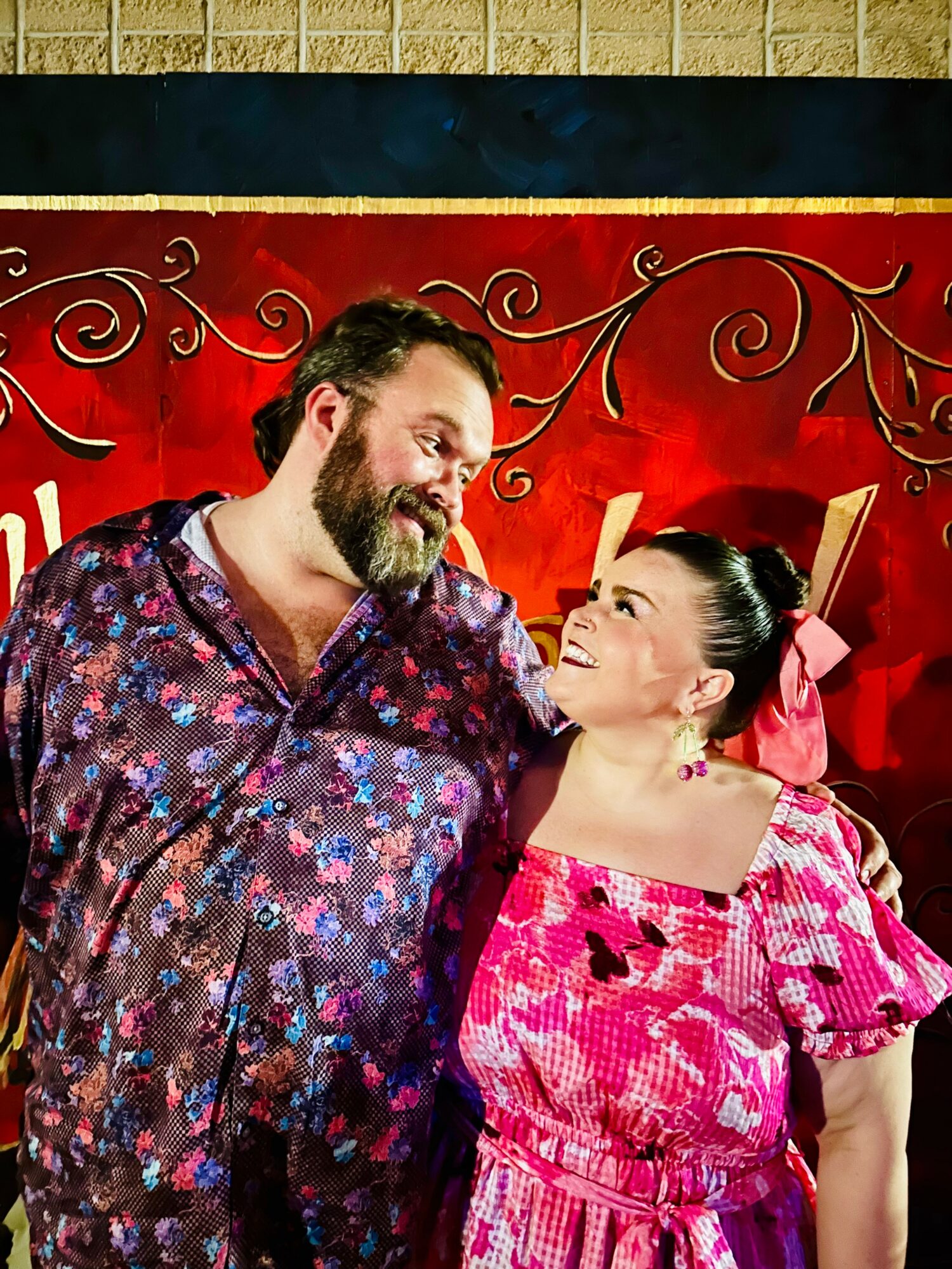 Two people smiling and looking at each other, standing in front of a red decorative background with swirls.
