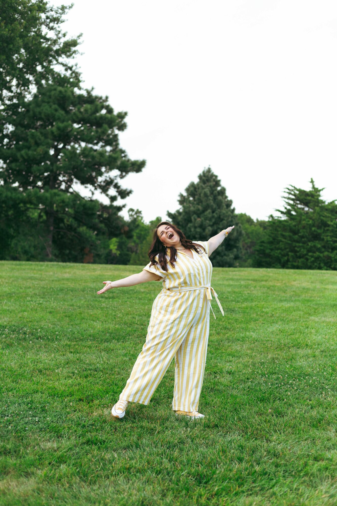 Woman in yellow and white striped jumpsuit standing on grass with arms outstretched, trees in background