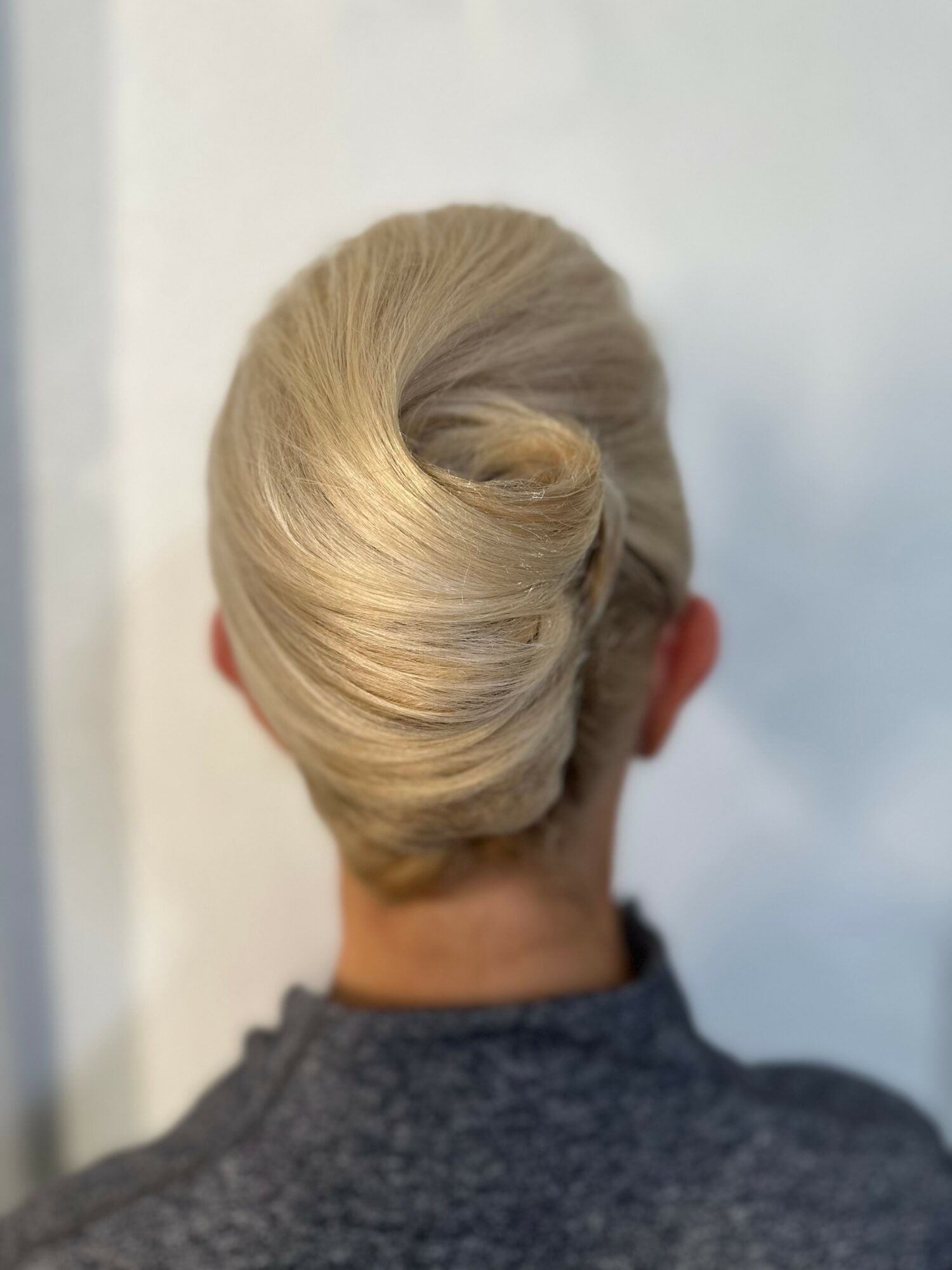 Elegant blonde updo hairstyle with a rolled bun at the back of the head, viewed from behind, with a gray top.