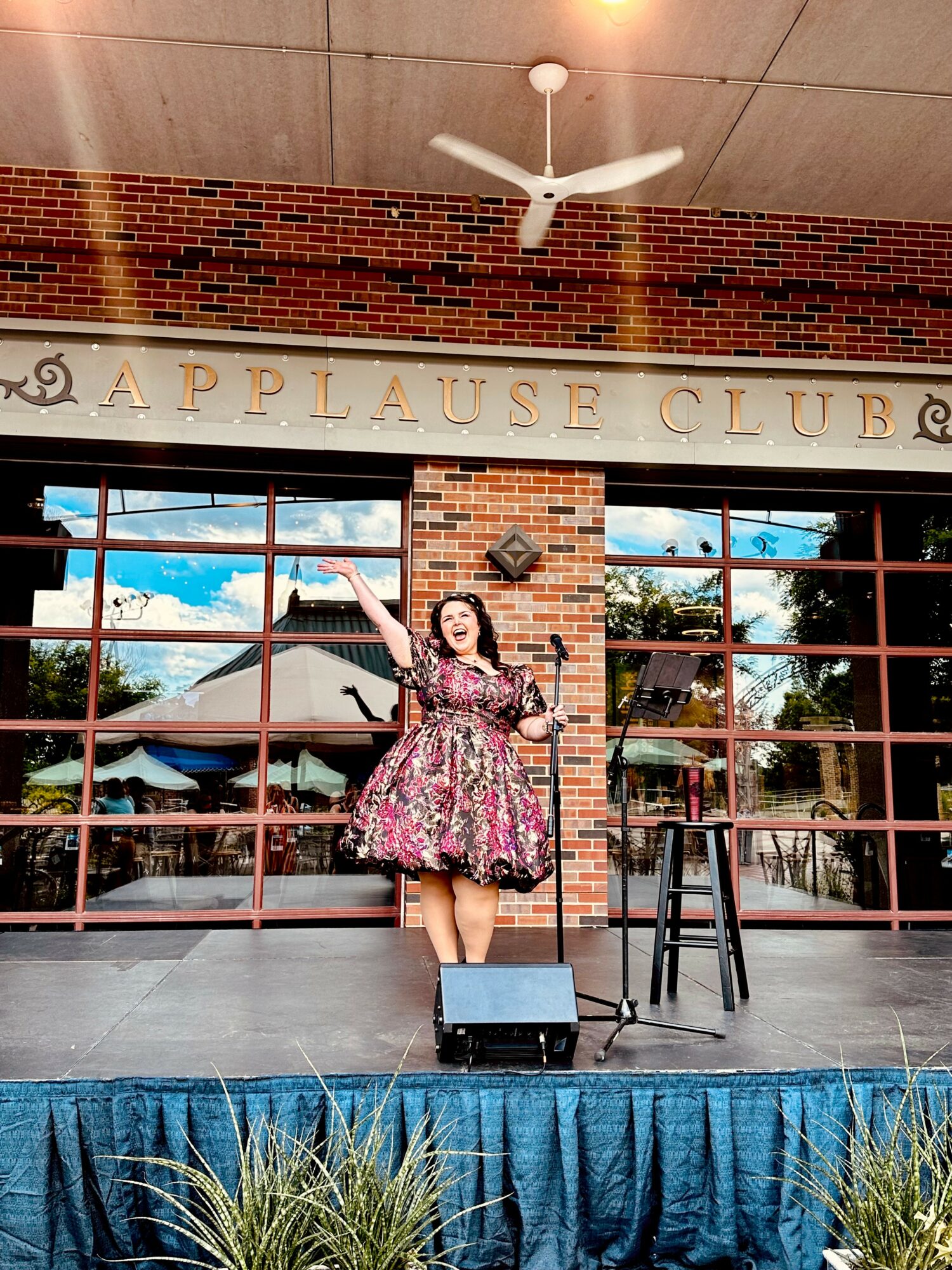 Woman in floral dress on stage with brick wall and windows behind her, pointing upward, in an applause club setting.