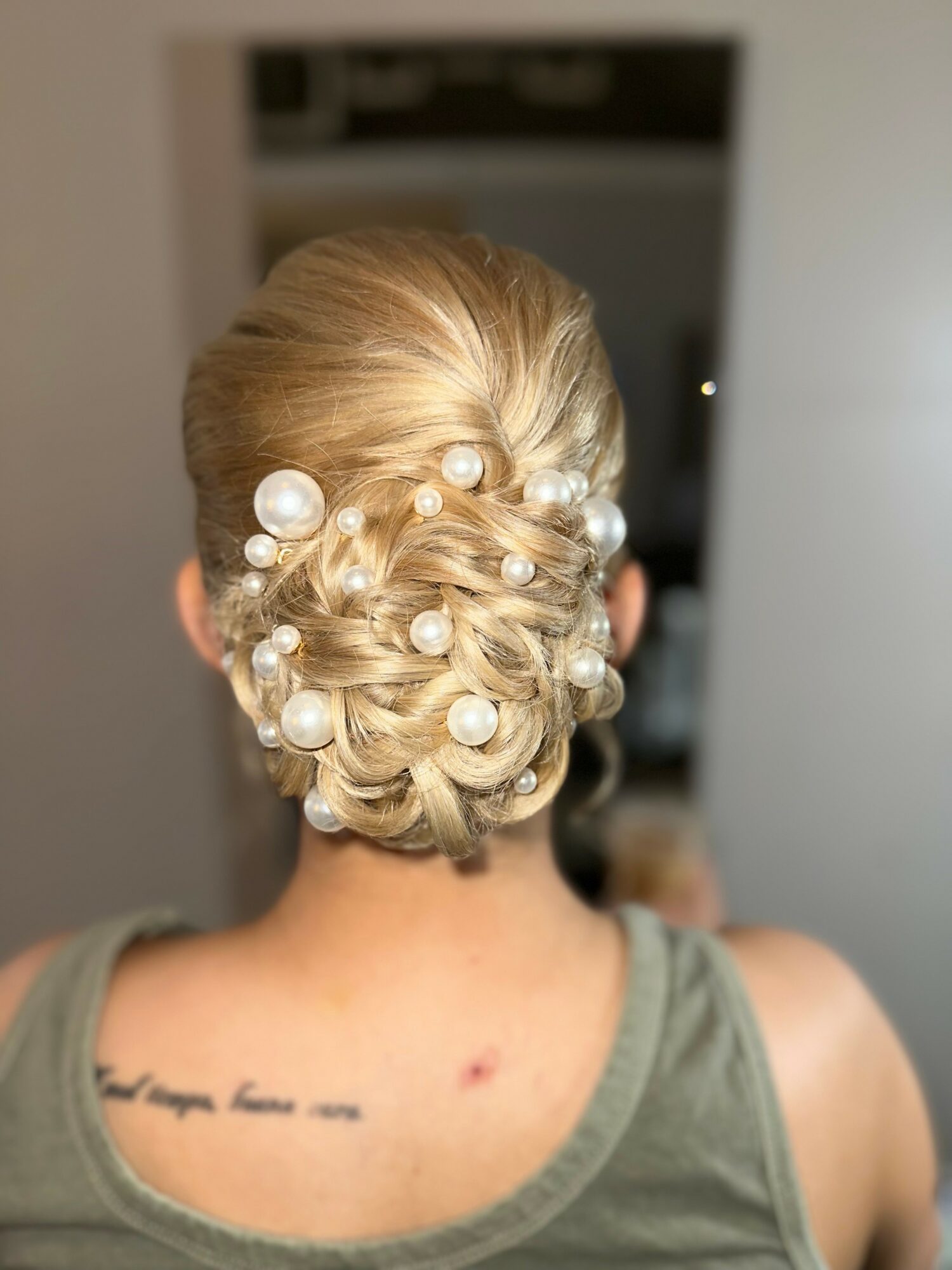 Back of a woman's head with an elegant updo decorated with large pearls, wearing a green tank top.