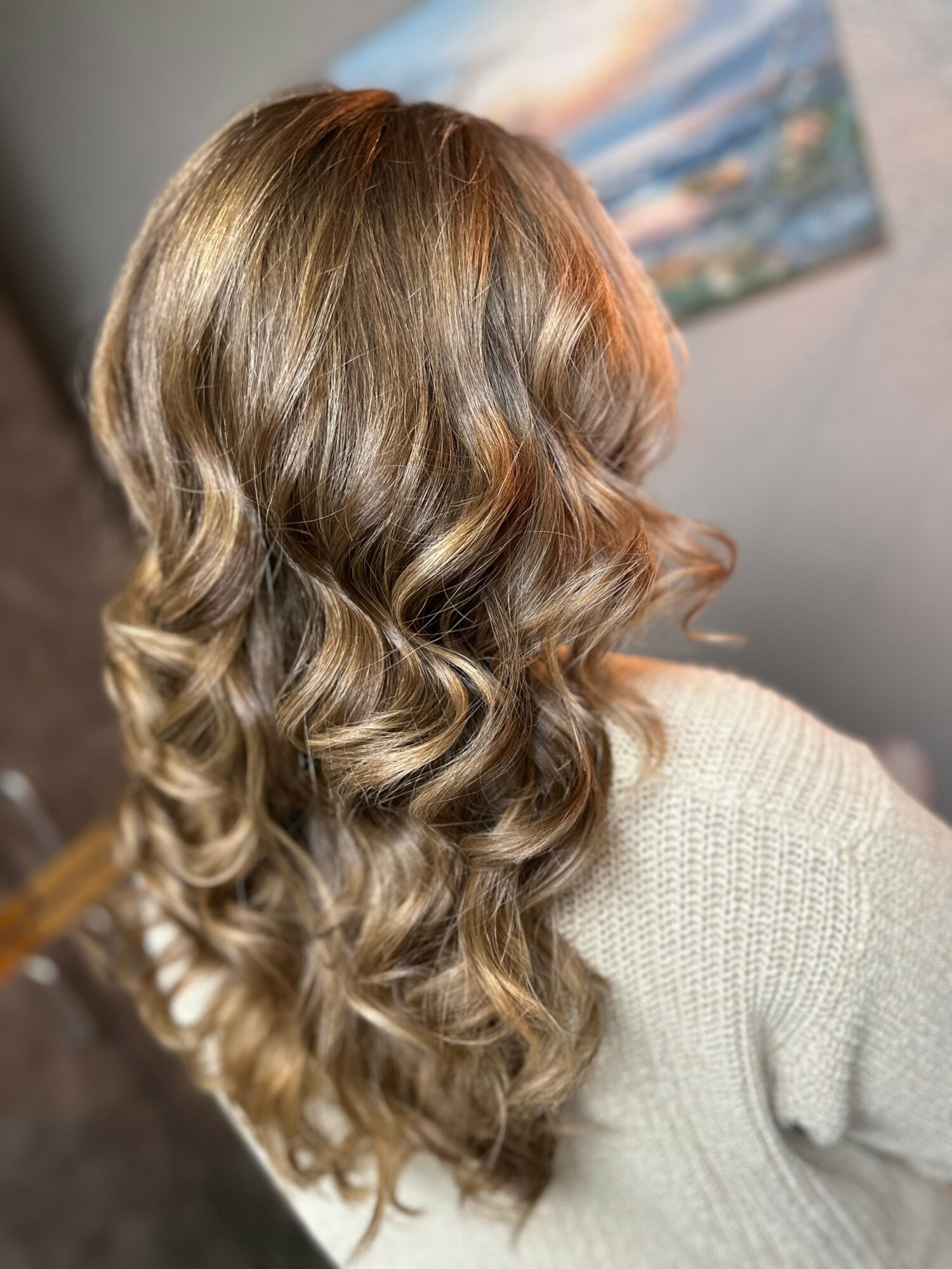 Back of a person's head with long, wavy, blonde hair, wearing a light-colored sweater, with a blurred painting in the background.