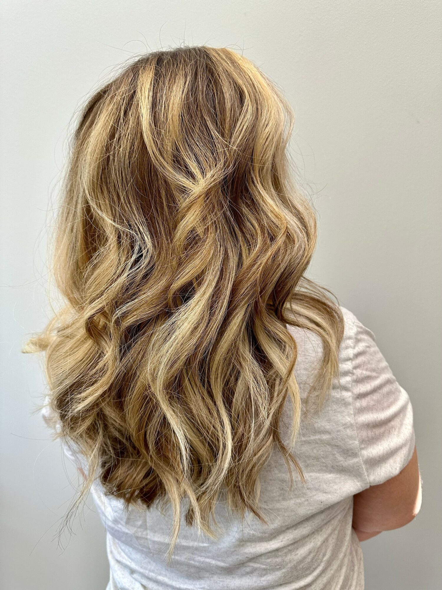 Back view of a woman with long, wavy blonde hair wearing a light-colored shirt.