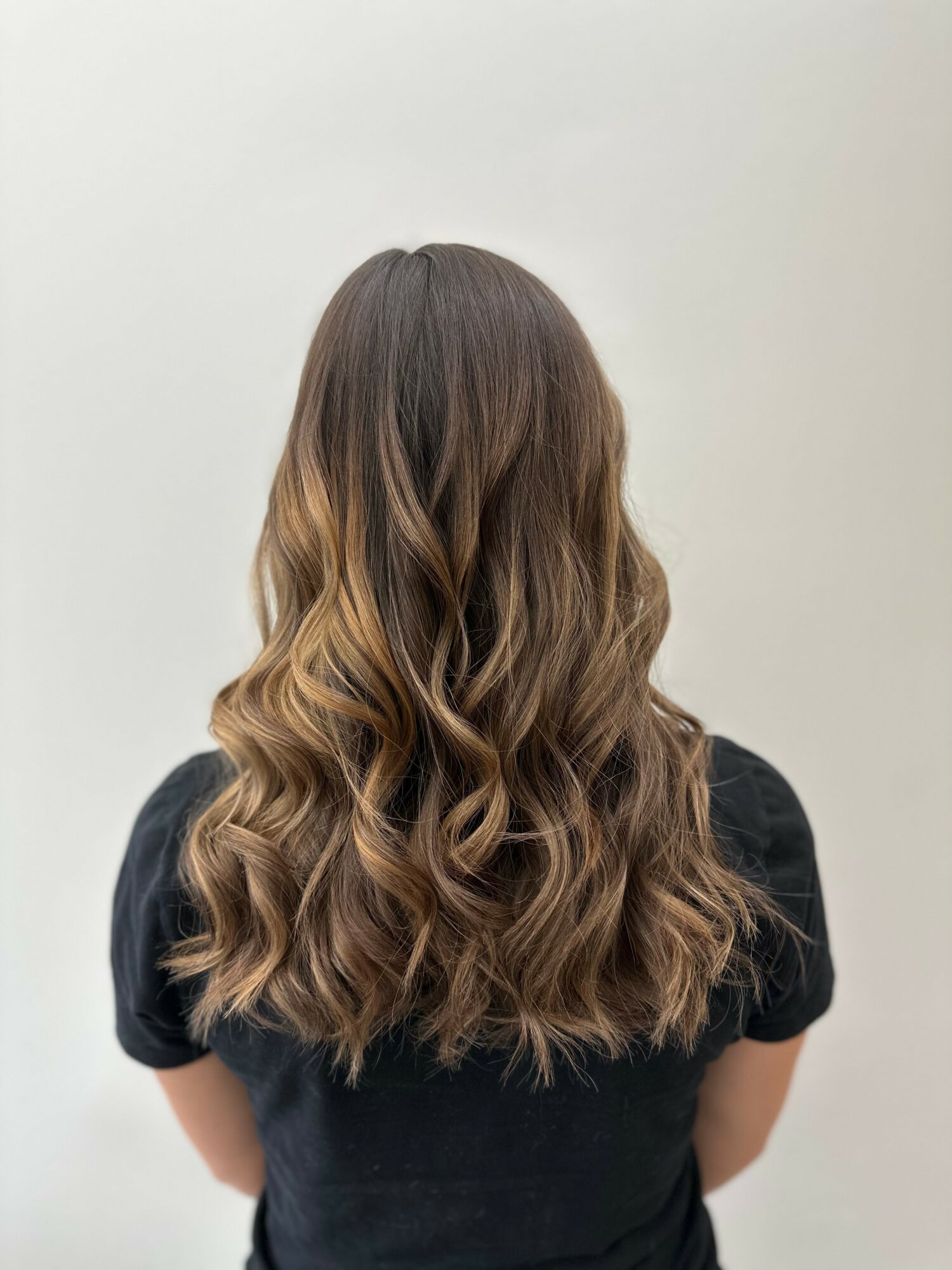 Back view of a person with long, wavy brown hair wearing a black top, standing against a plain background.