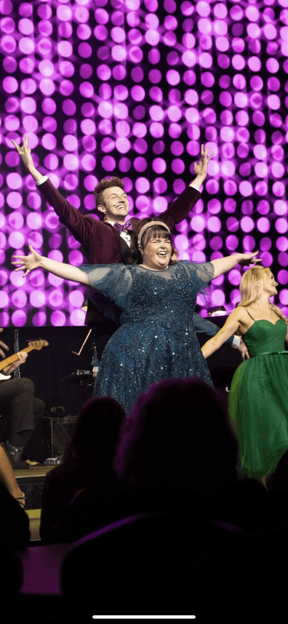 Two performers singing on stage with a purple dotted background, one in a blue dress and the other in a green dress.
