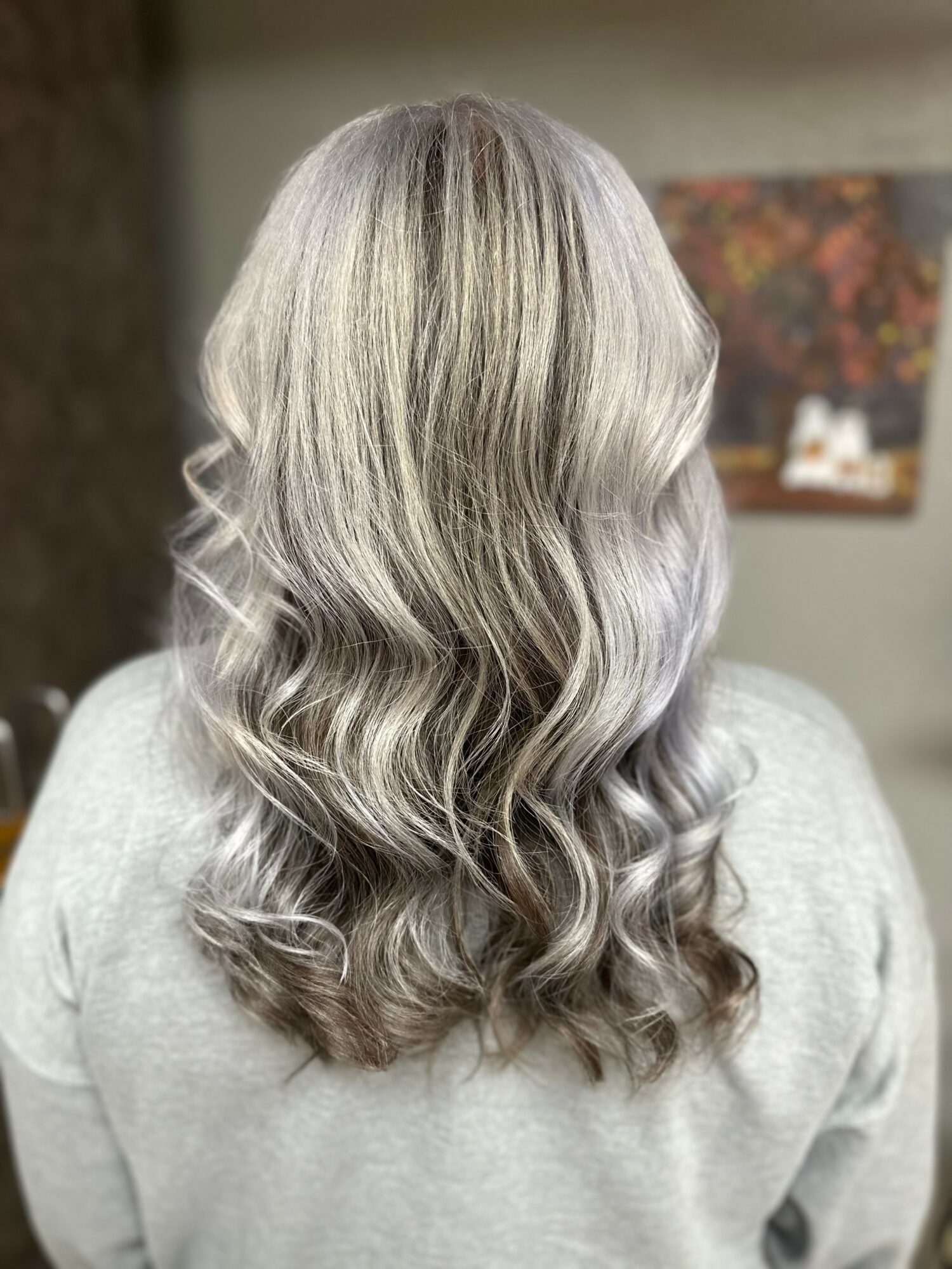 Back view of a person with long, wavy, silver-gray hair, wearing a light-colored top, in an indoor setting.