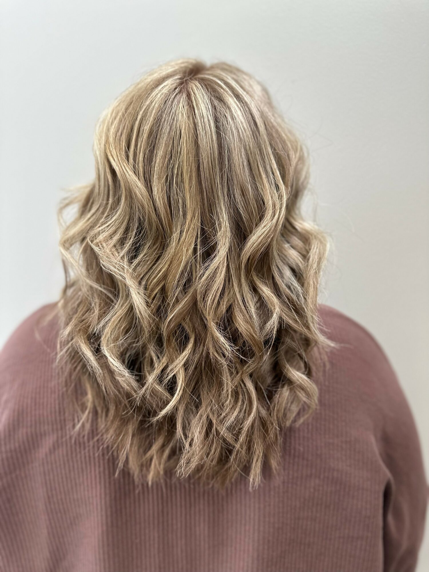 Back view of a person with long, wavy blonde hair wearing a brown top, against a plain light background.