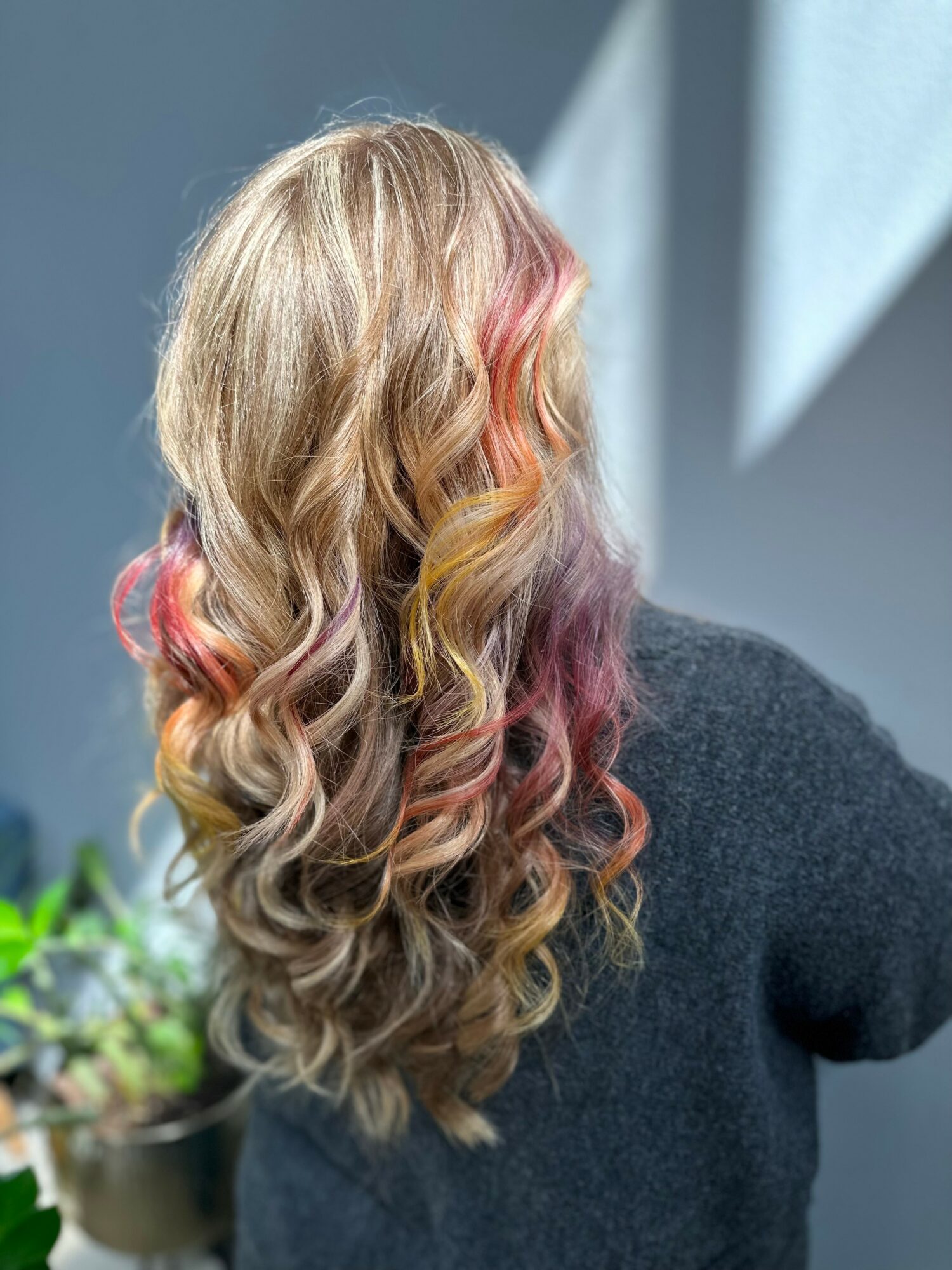 Back view of a person with long, wavy hair featuring blonde, pink, orange, and purple colors, wearing a dark sweater.