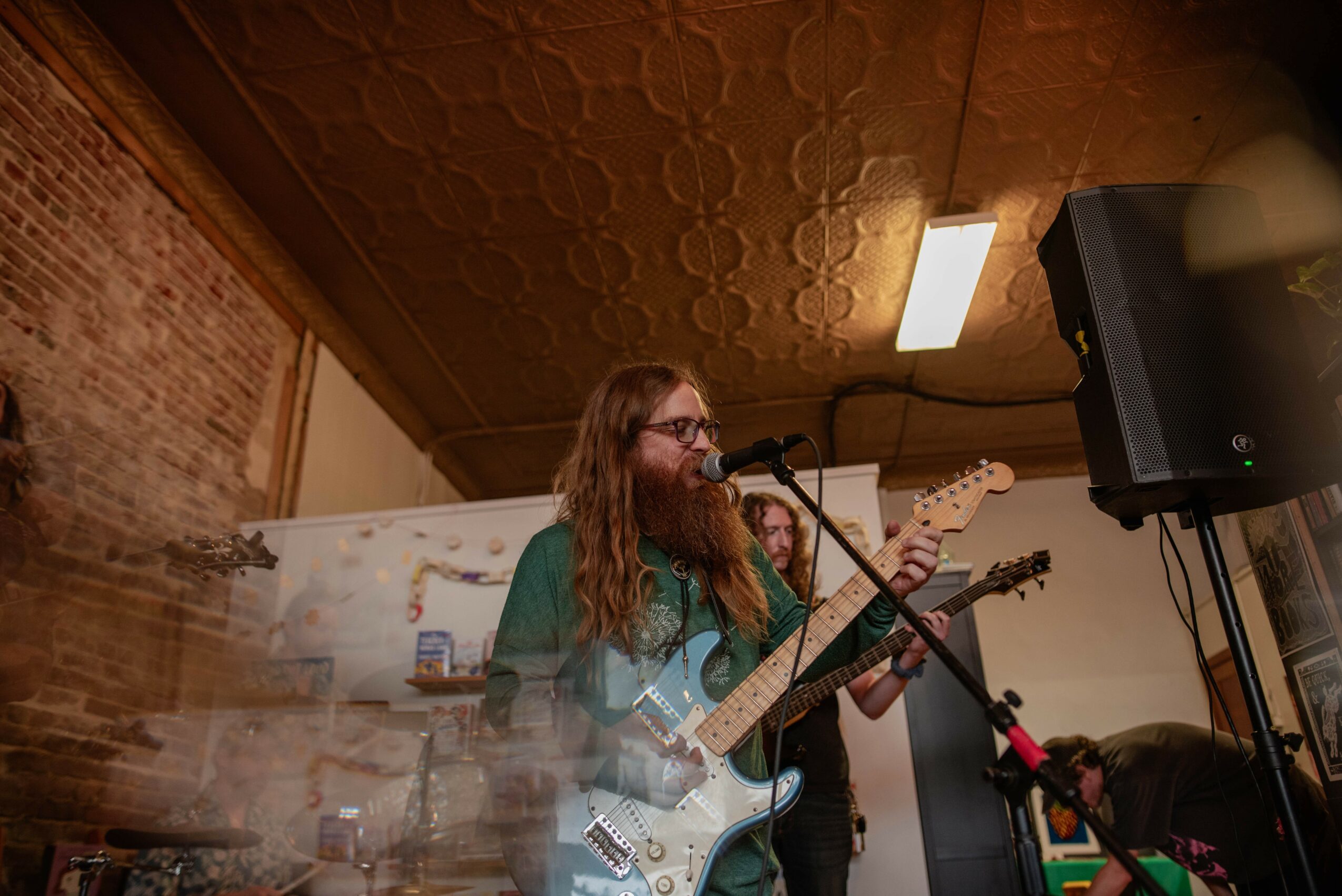 Person with long hair and glasses playing an electric guitar and singing into a microphone indoors.