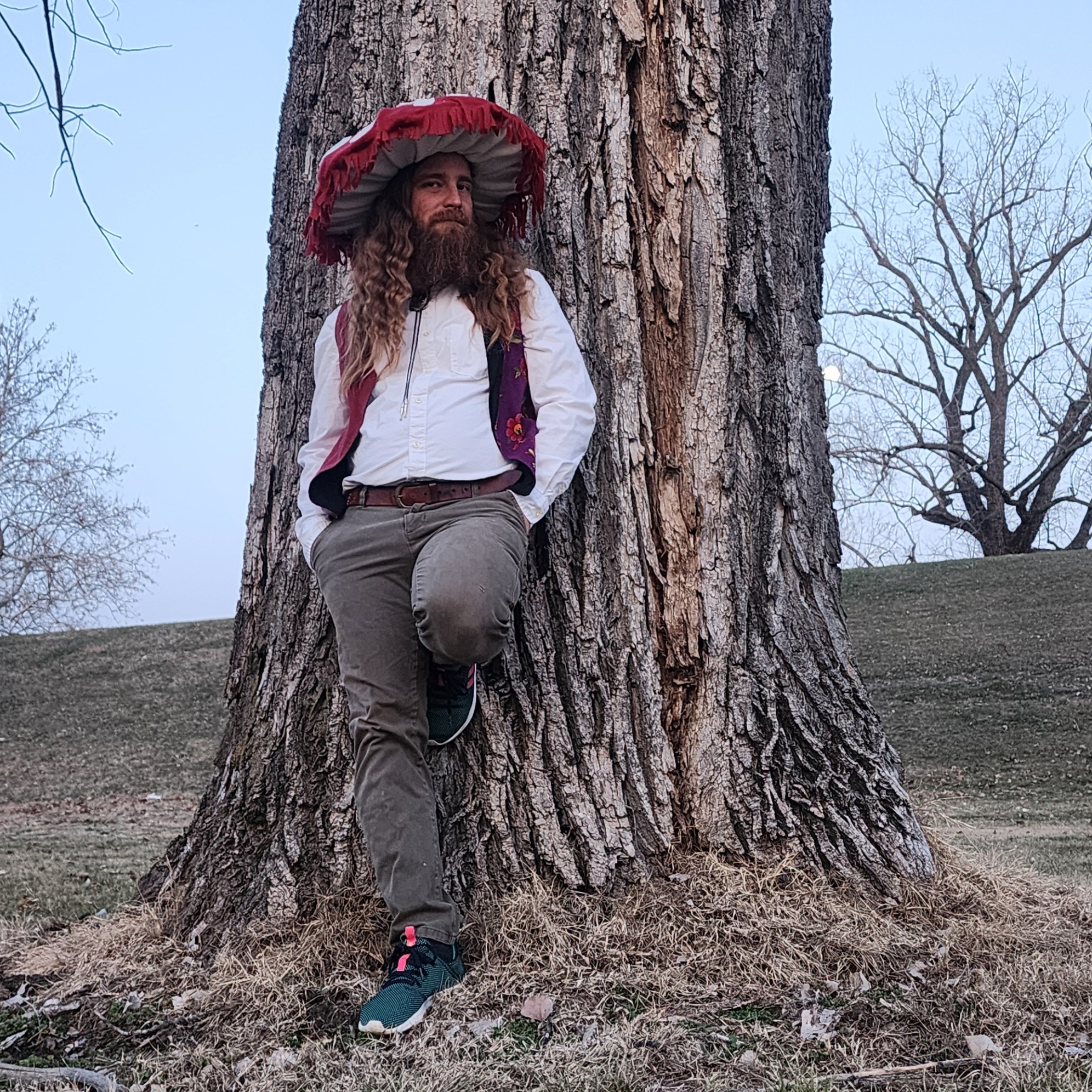 Person with long hair and beard leaning against a large tree outdoors, wearing a red hat, white shirt, and gray pants.