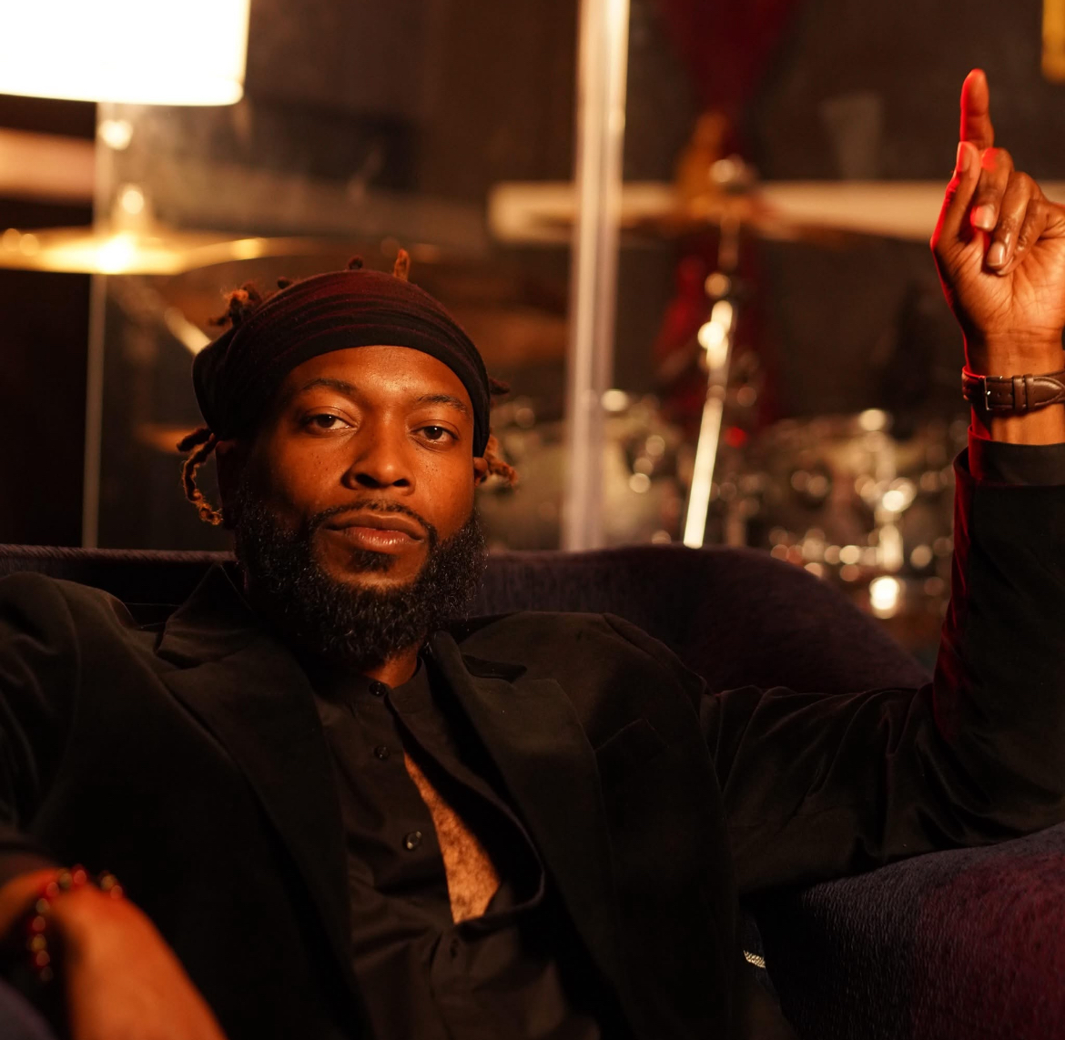 Man with a beard and headband sitting on a dark sofa, raising his right hand with one finger pointing up, in a dimly lit room.