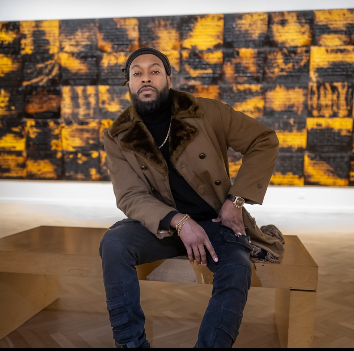 Man sitting on a wooden bench in front of an abstract yellow and black artwork, wearing a brown jacket and black pants.