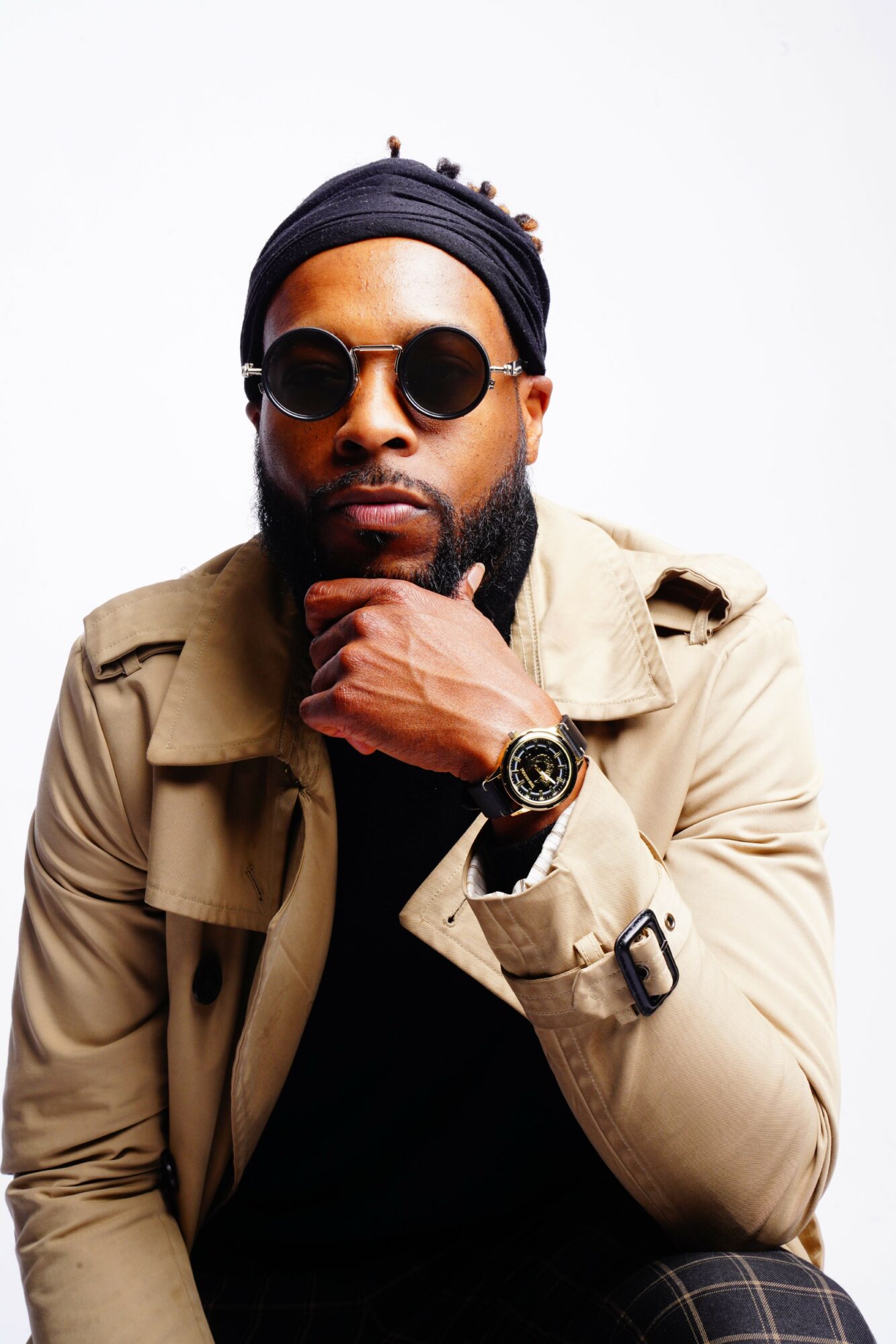 Man with sunglasses and a watch, wearing a beige jacket, poses with hand near chin against white background.