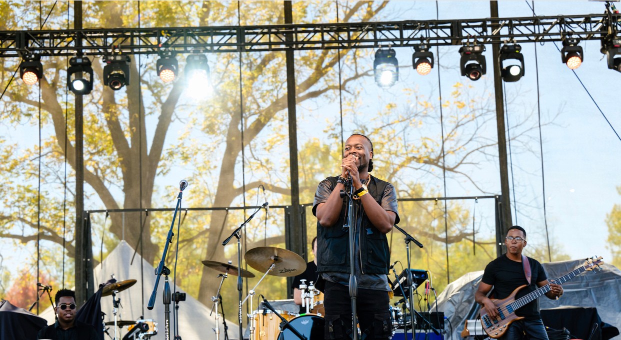 Band performing on outdoor stage with trees and sunlight in background, singer at center holding microphone, musicians playing instruments.