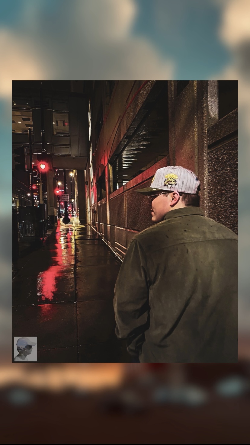 Person wearing a cap and jacket walking on a wet city street at night with reflections and red traffic lights.