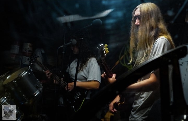 Two musicians playing guitars and drums in a dark, dimly lit setting, with microphones and musical equipment visible.