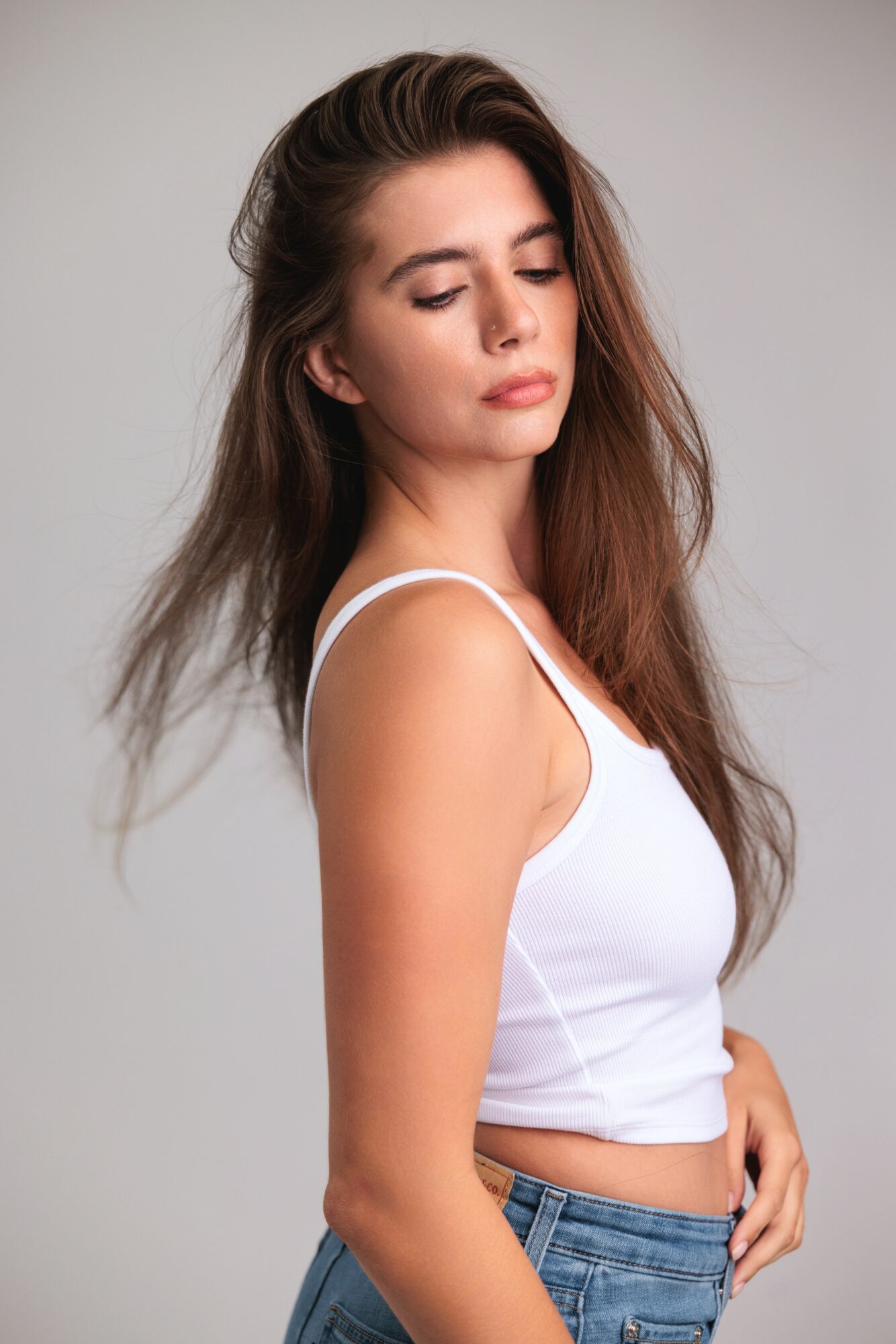 Young woman with long brown hair wearing a white tank top and blue jeans, looking down with eyes closed.