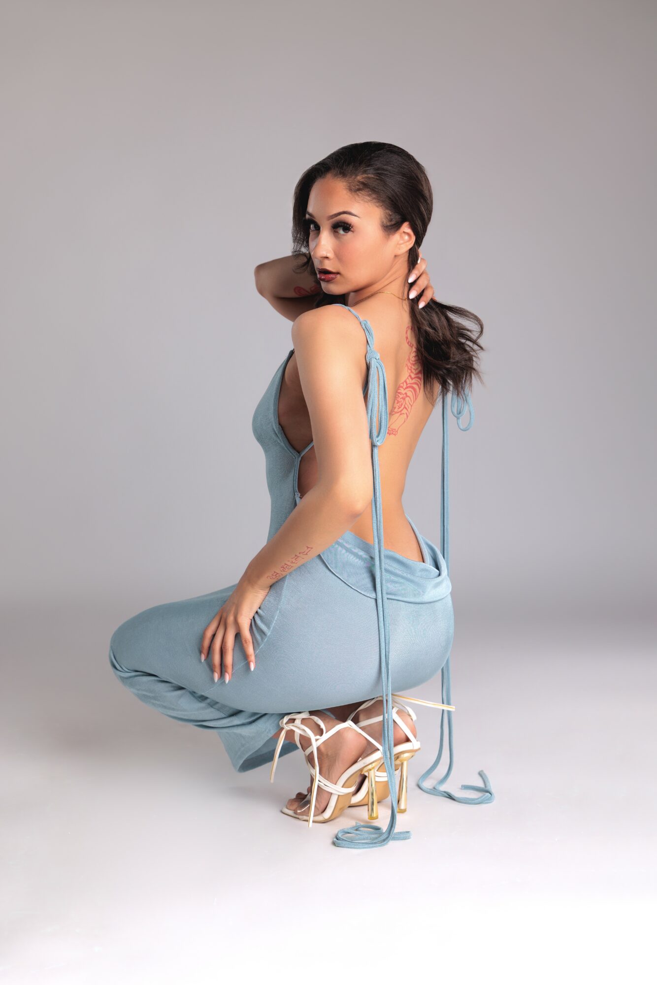 Woman in light blue dress crouching with one hand on her knee and the other touching her hair, against a plain background.