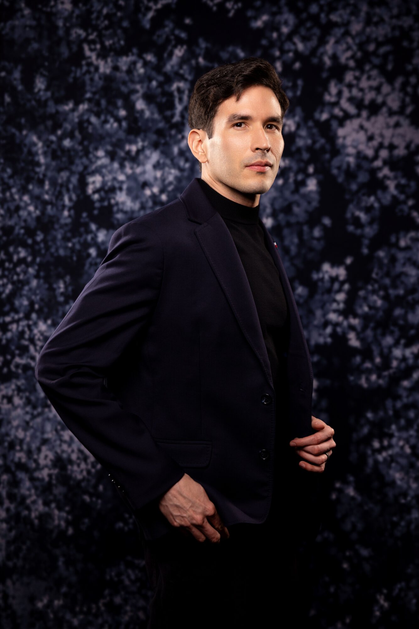 Man in black suit and turtleneck against a dark, textured background, looking to the side.