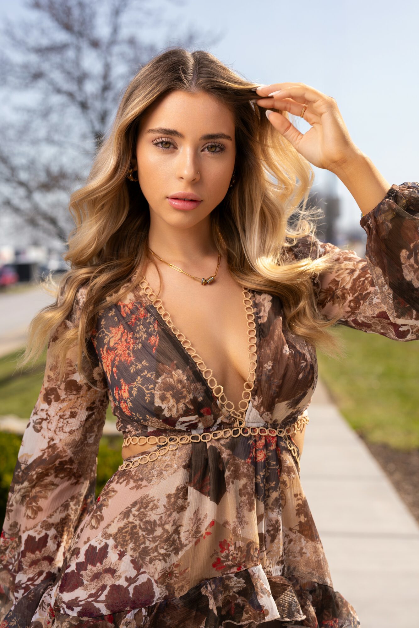 Young woman with wavy hair outdoors, wearing a patterned dress, looking at the camera, with trees and a sidewalk in background.